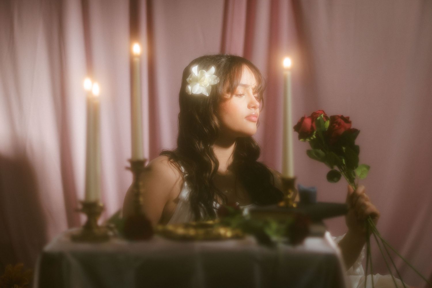 Serene woman with floral hair accessory holding red roses at elegant candlelit table setting.