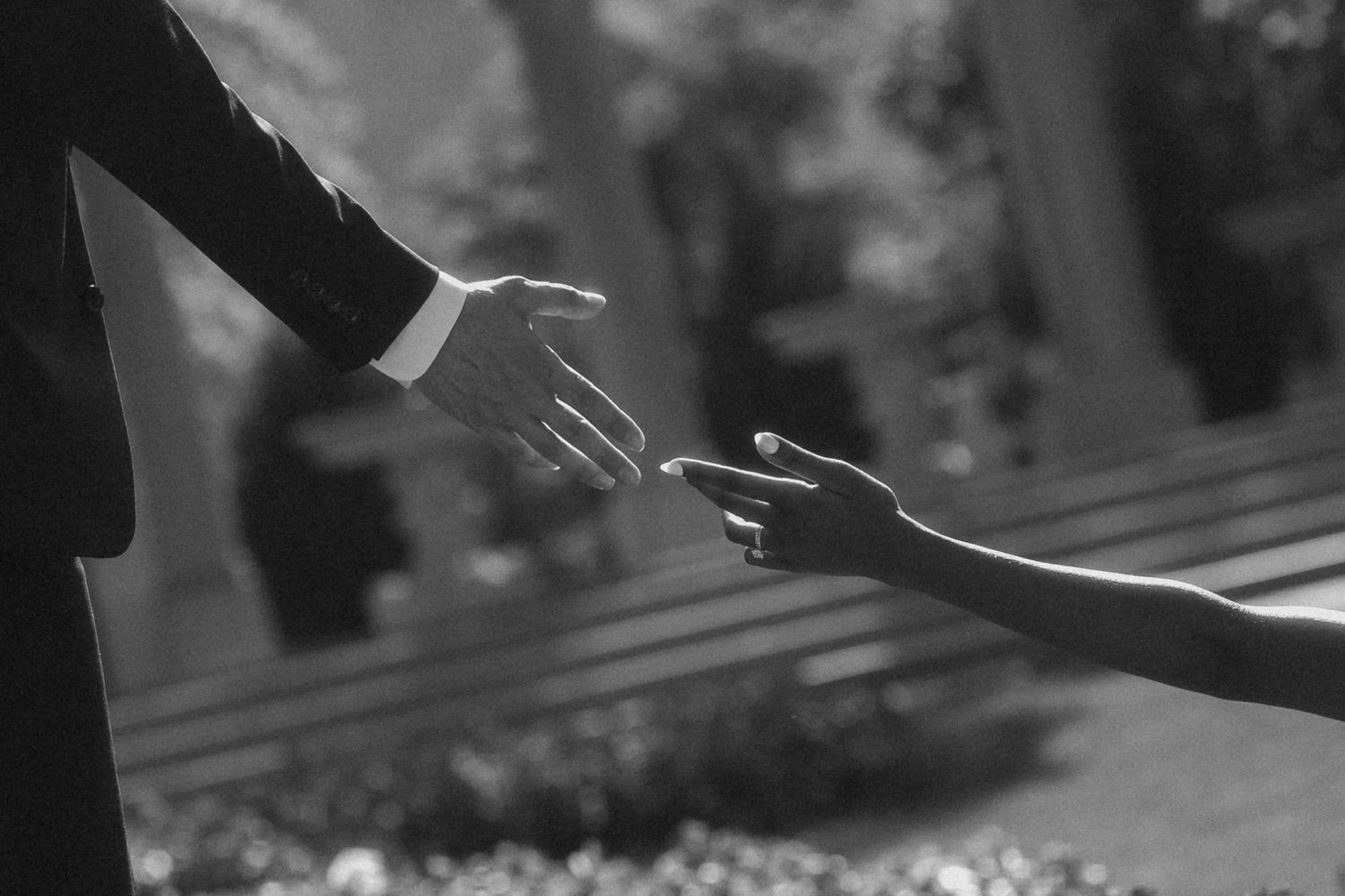 Black and white photo of two hands reaching toward each other, one wearing an engagement ring, in a romantic outdoor setting.