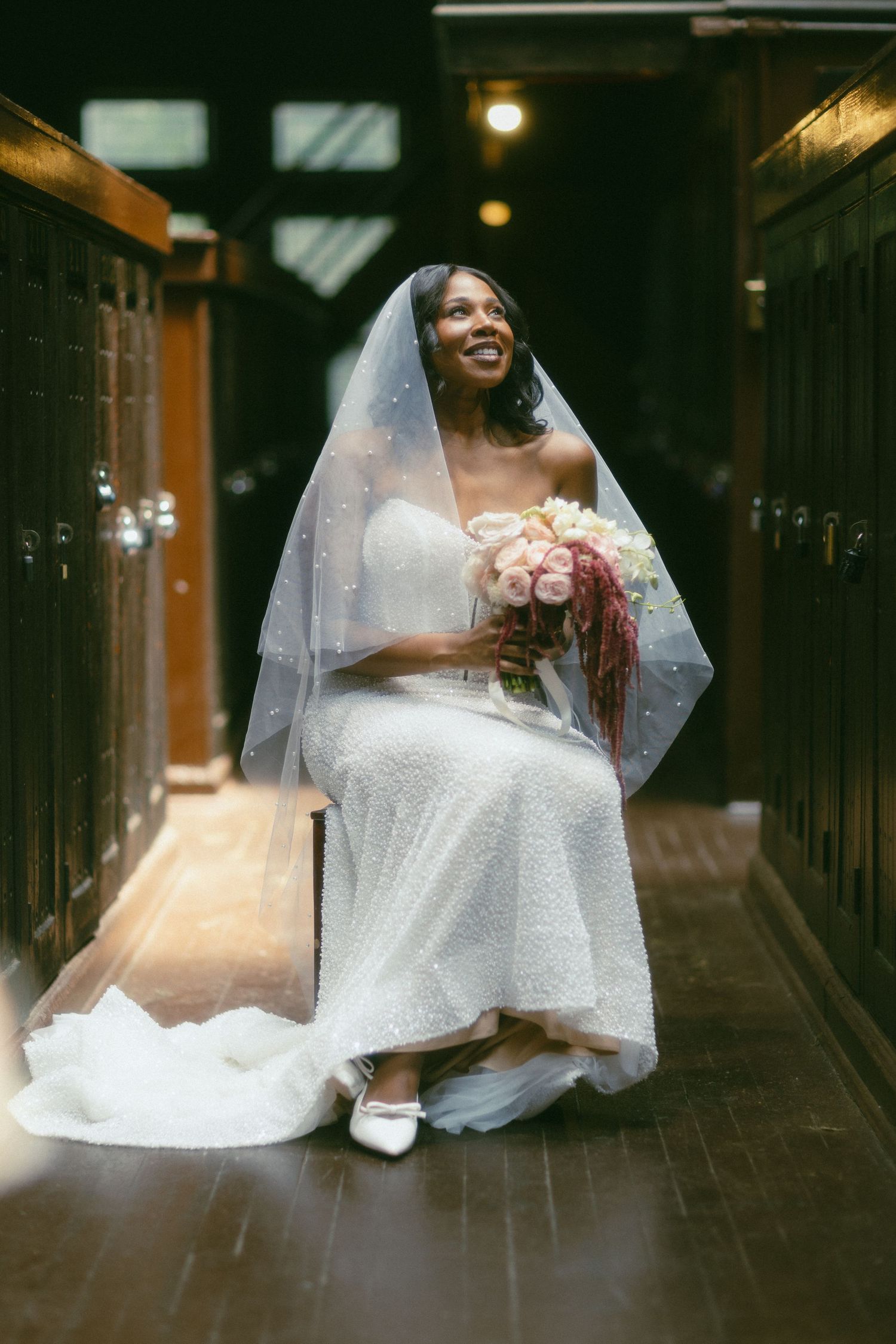Radiant bride in white beaded gown and veil sits smiling holding pink bouquet in wood-paneled hall.