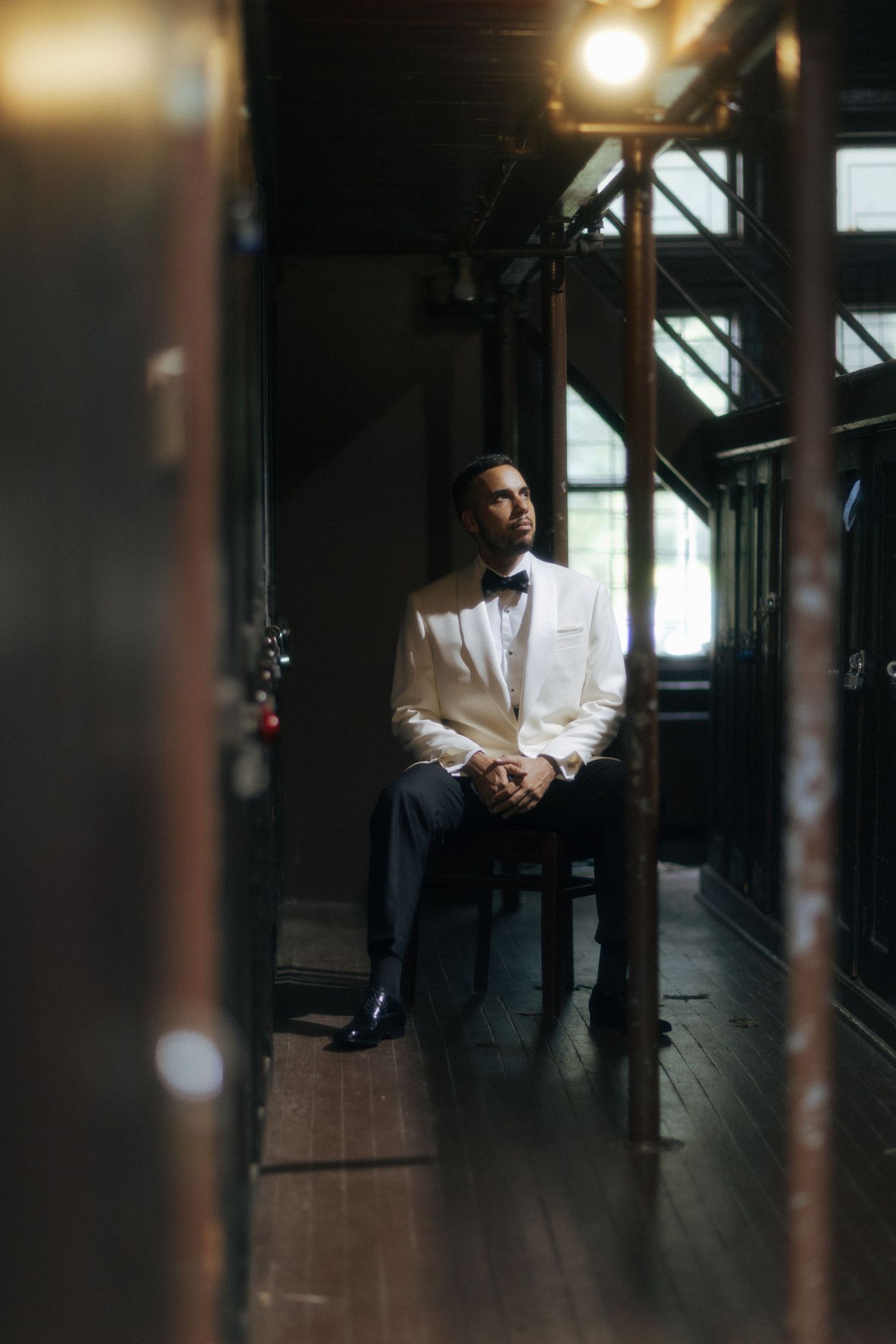 Groom in white tuxedo jacket and bow tie sits pensively in dimly lit elegant venue before wedding ceremony.