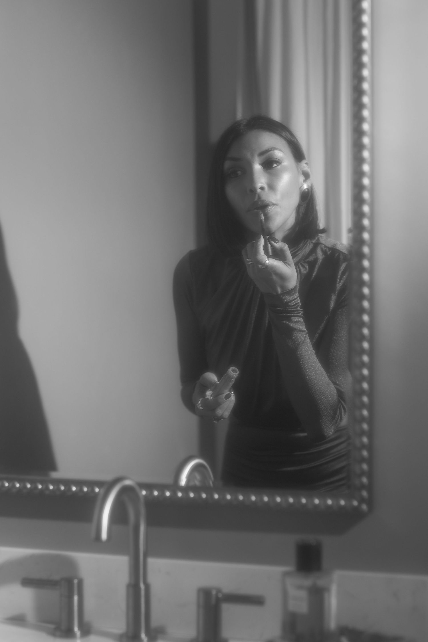 Black and white photo of a woman applying lipstick while looking in a mirror above a bathroom sink.