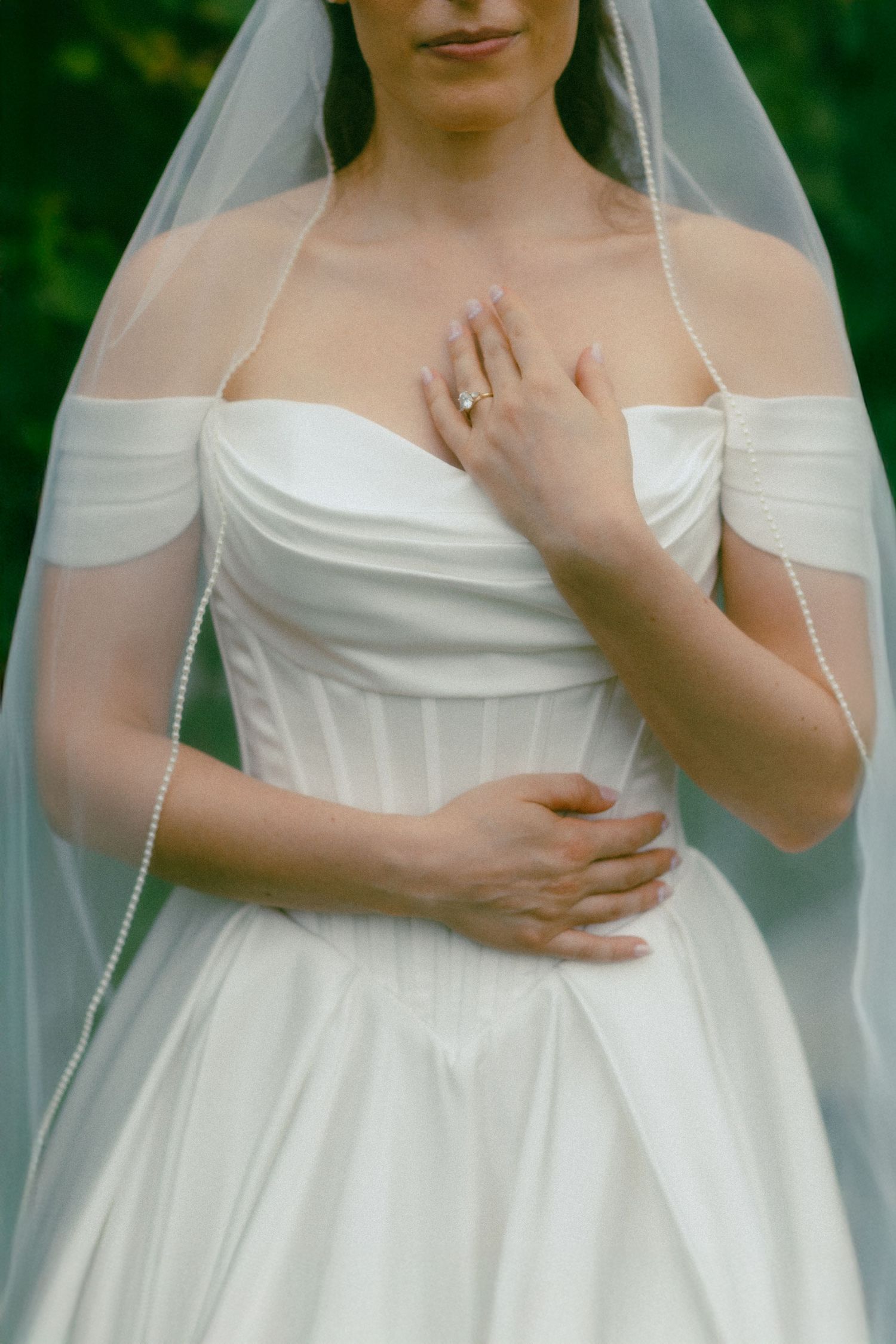 Bride in off-shoulder white gown and pearl-trimmed veil, showcasing engagement ring with hands crossed over chest.