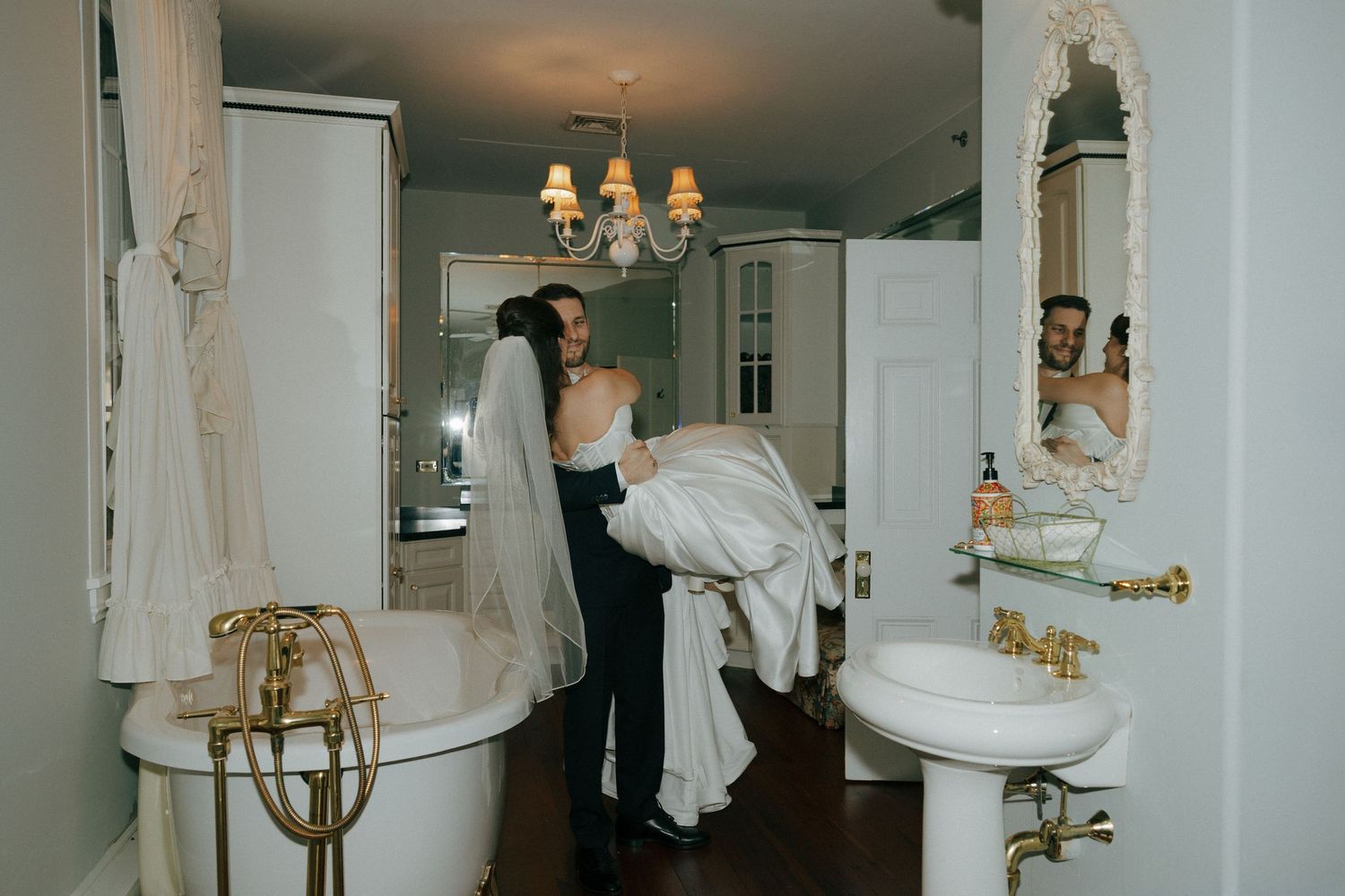 Groom helping bride with wedding dress in luxurious vintage bathroom with clawfoot tub and gold fixtures.