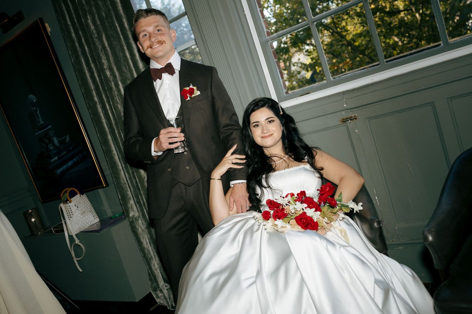 Bride in white ballgown and groom in brown tuxedo pose elegantly indoors with red roses.