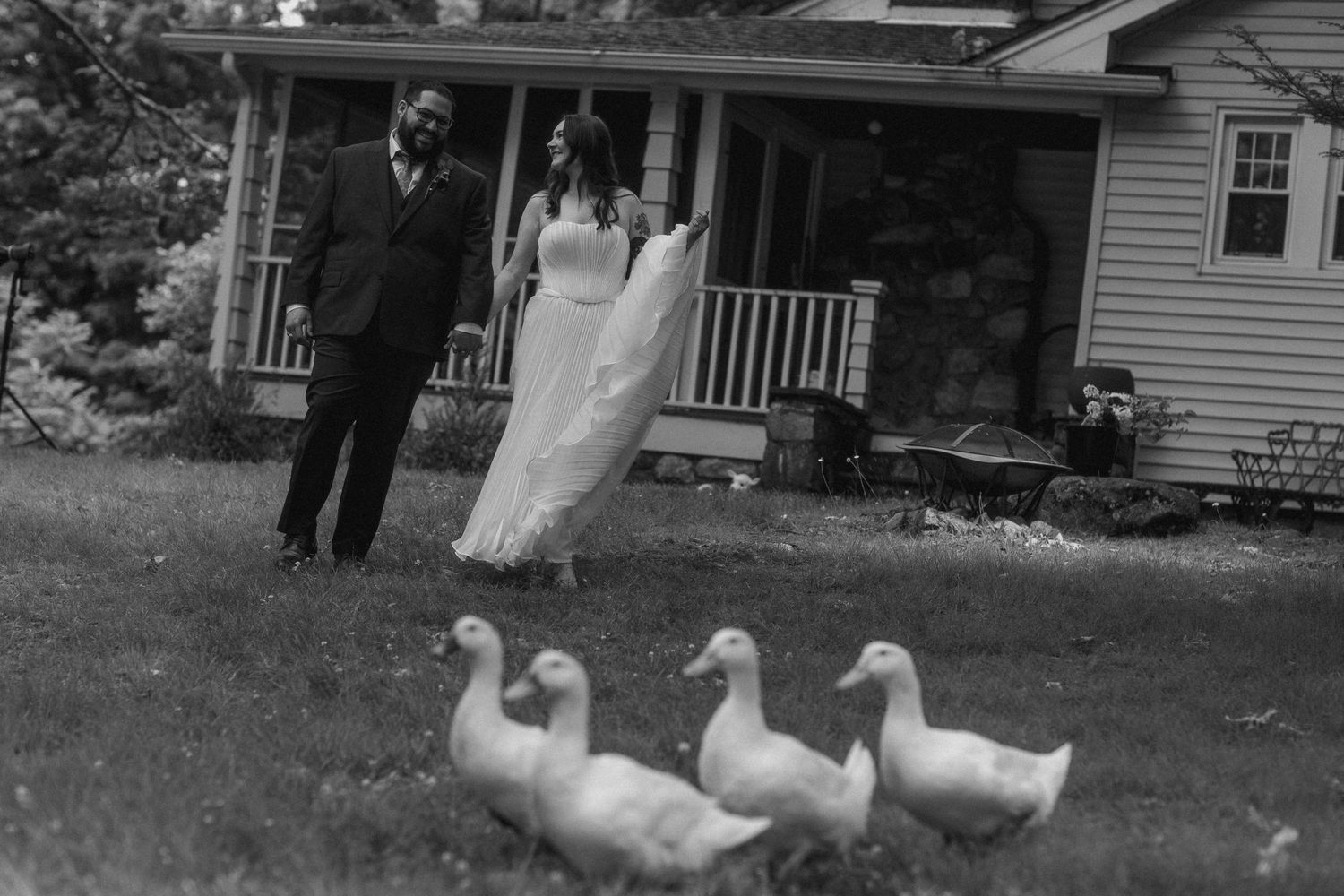 Black and white photo of a bride and groom walking near three white ducks on a lawn outside a house.