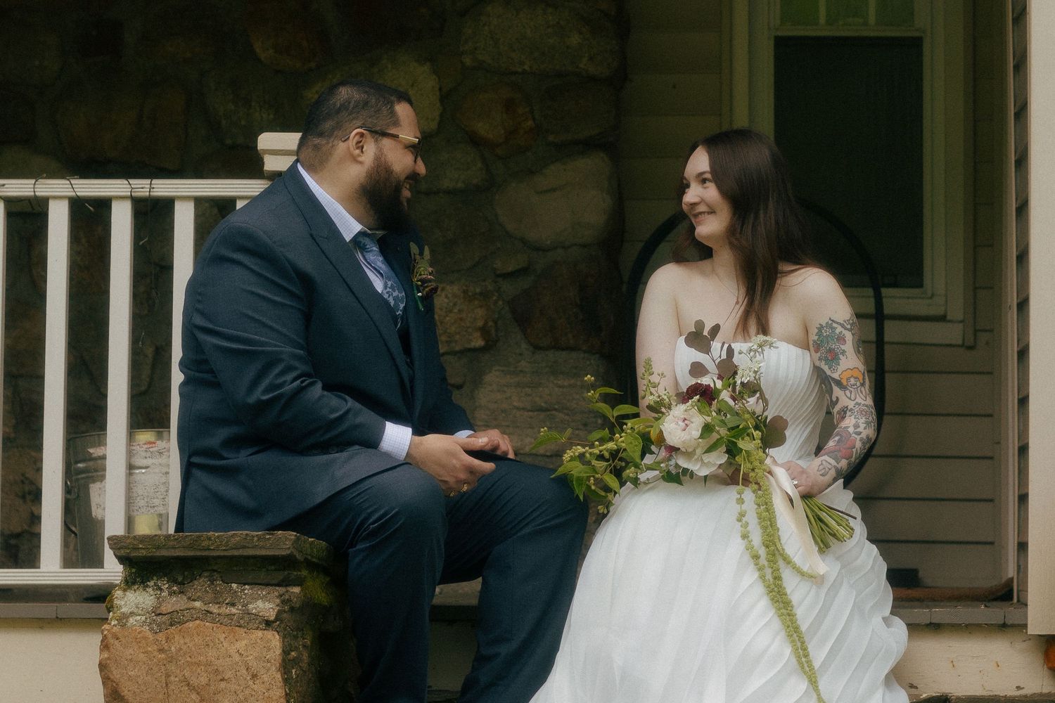 Bride and groom sitting together on stone steps, bride holding floral bouquet, groom in navy suit, sharing a joyful moment.
