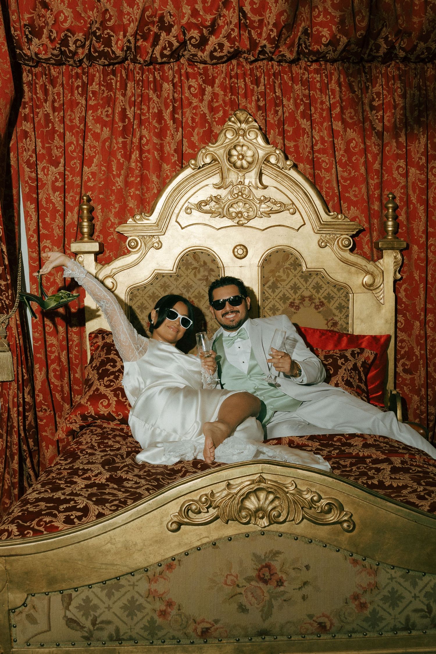 Couple in white robes and sunglasses lounging on ornate gold bed with red floral curtains, holding champagne glasses.