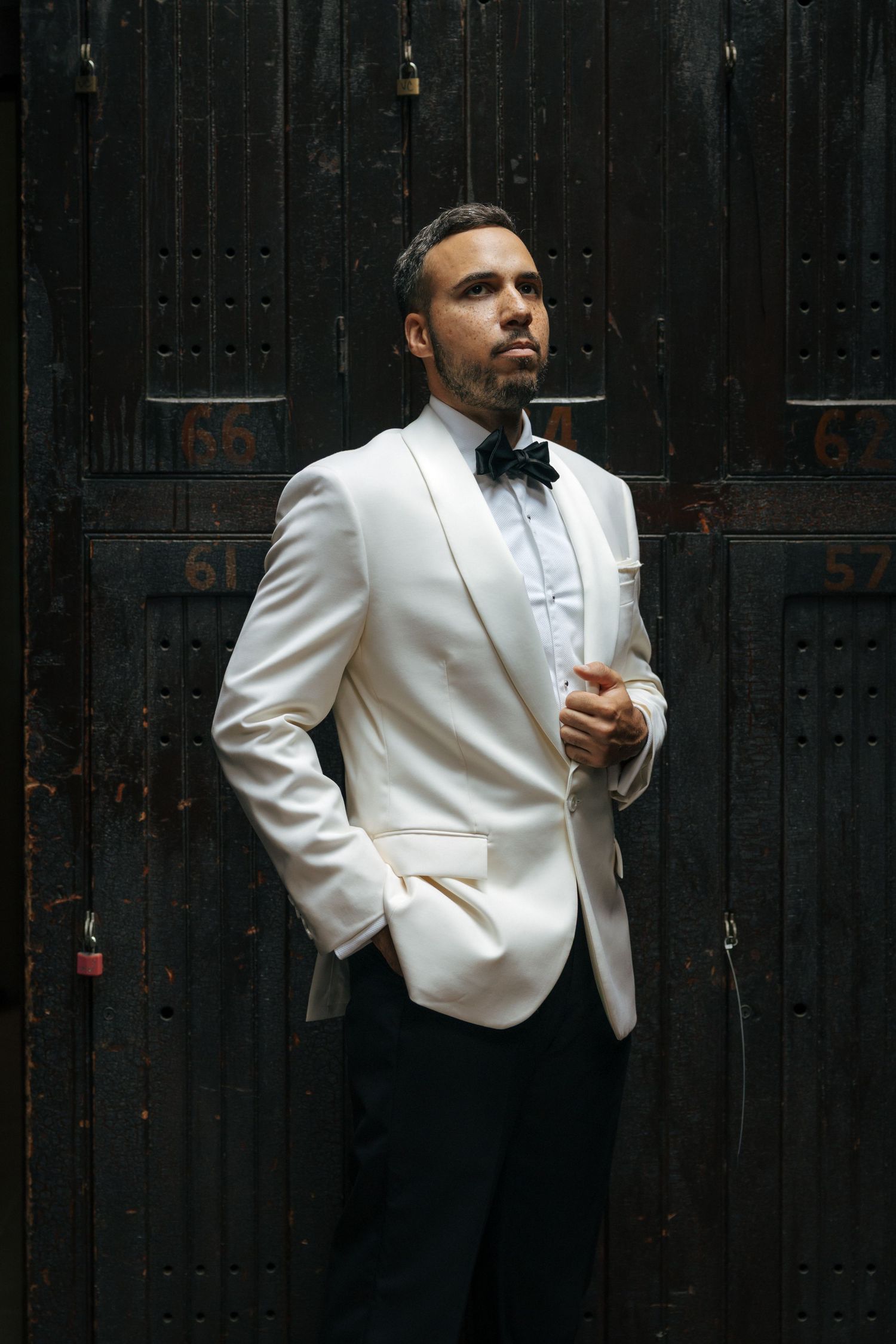 Elegant groom in white tuxedo jacket and black bow tie posing confidently against dark vintage doors.