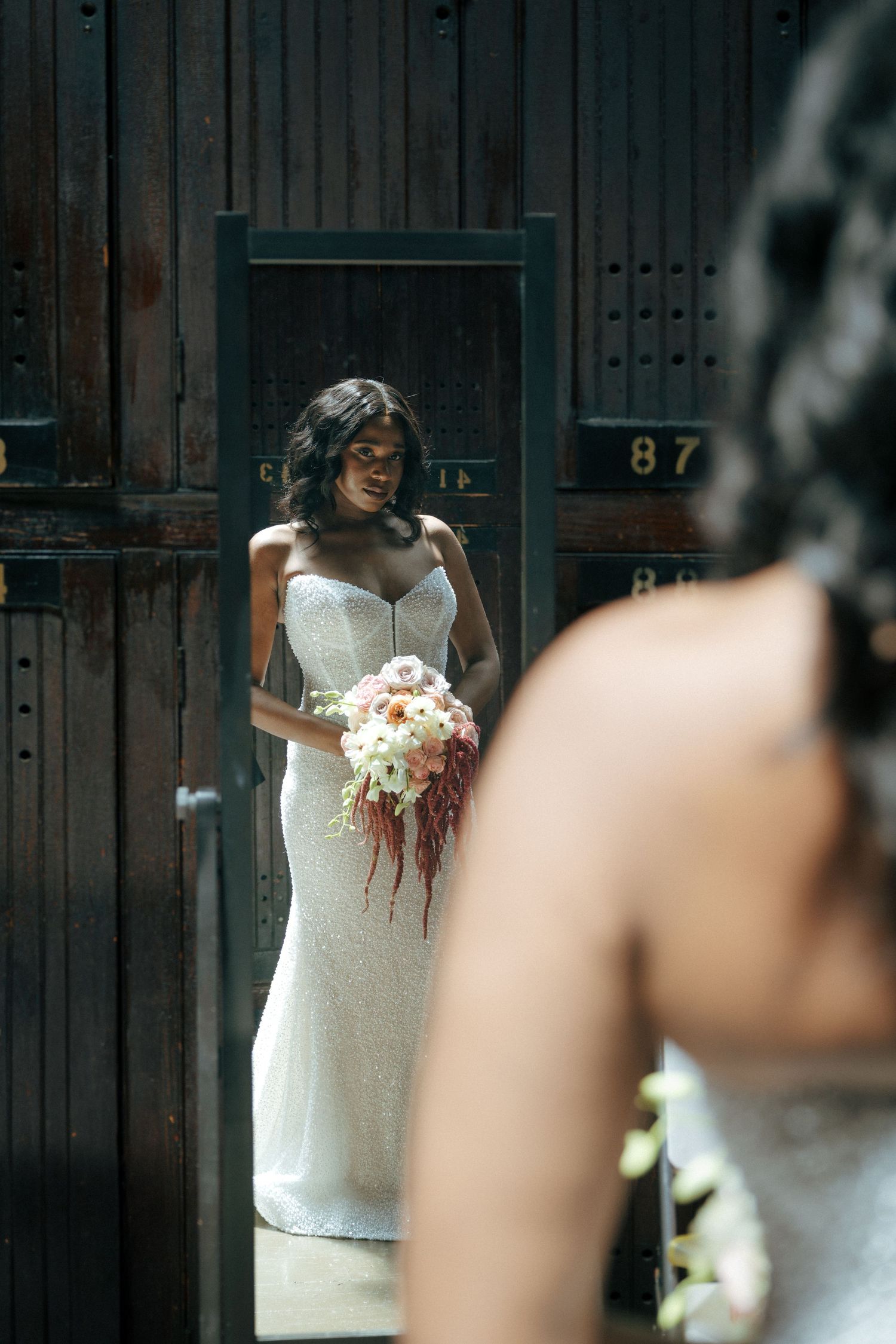 Bride in white sequin gown holding pink bouquet reflected in vintage mirror outdoors.