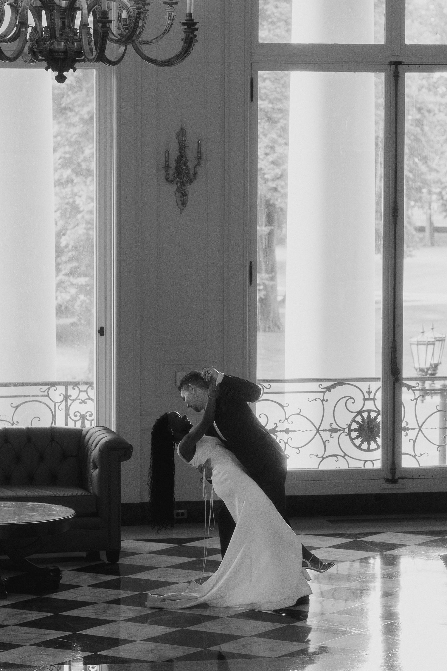 Black and white photo of a couple sharing a romantic dip kiss in an elegant ballroom with chandelier.