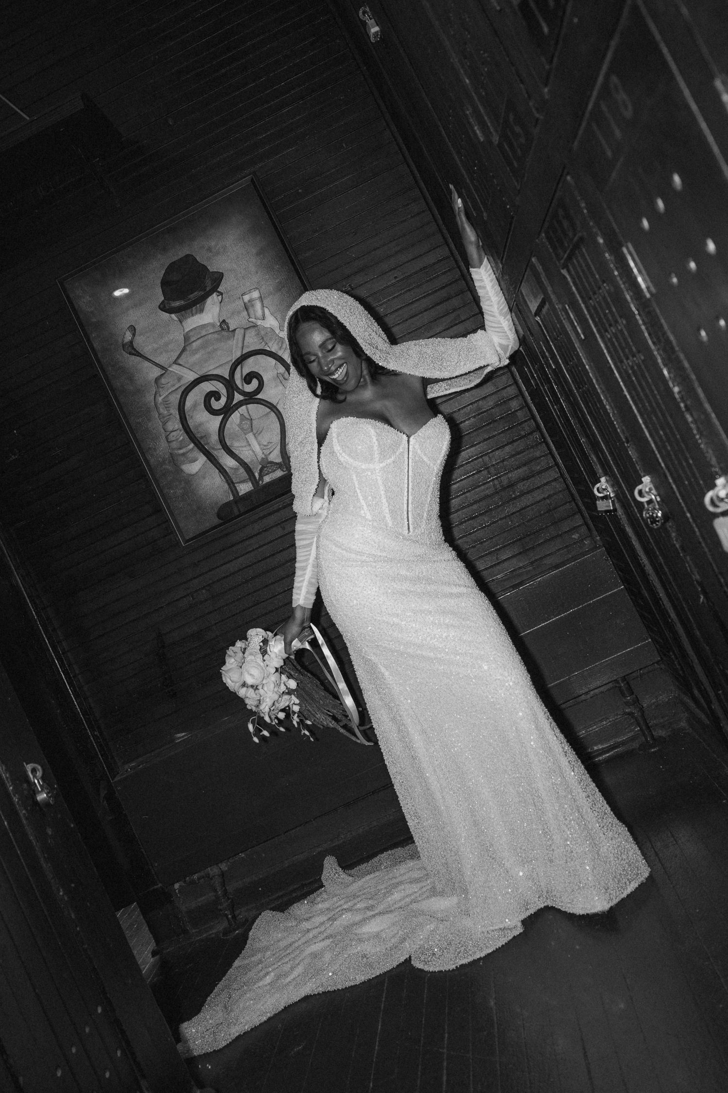 Black and white photo of a smiling bride in a sequined gown and hooded veil holding a bouquet indoors.