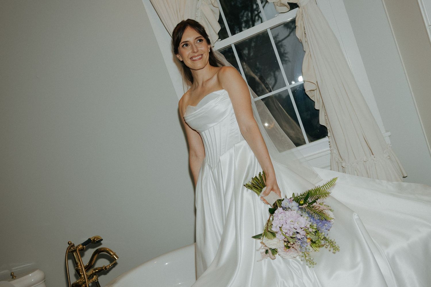 Smiling bride in strapless white satin wedding gown holding purple floral bouquet near window with curtains.