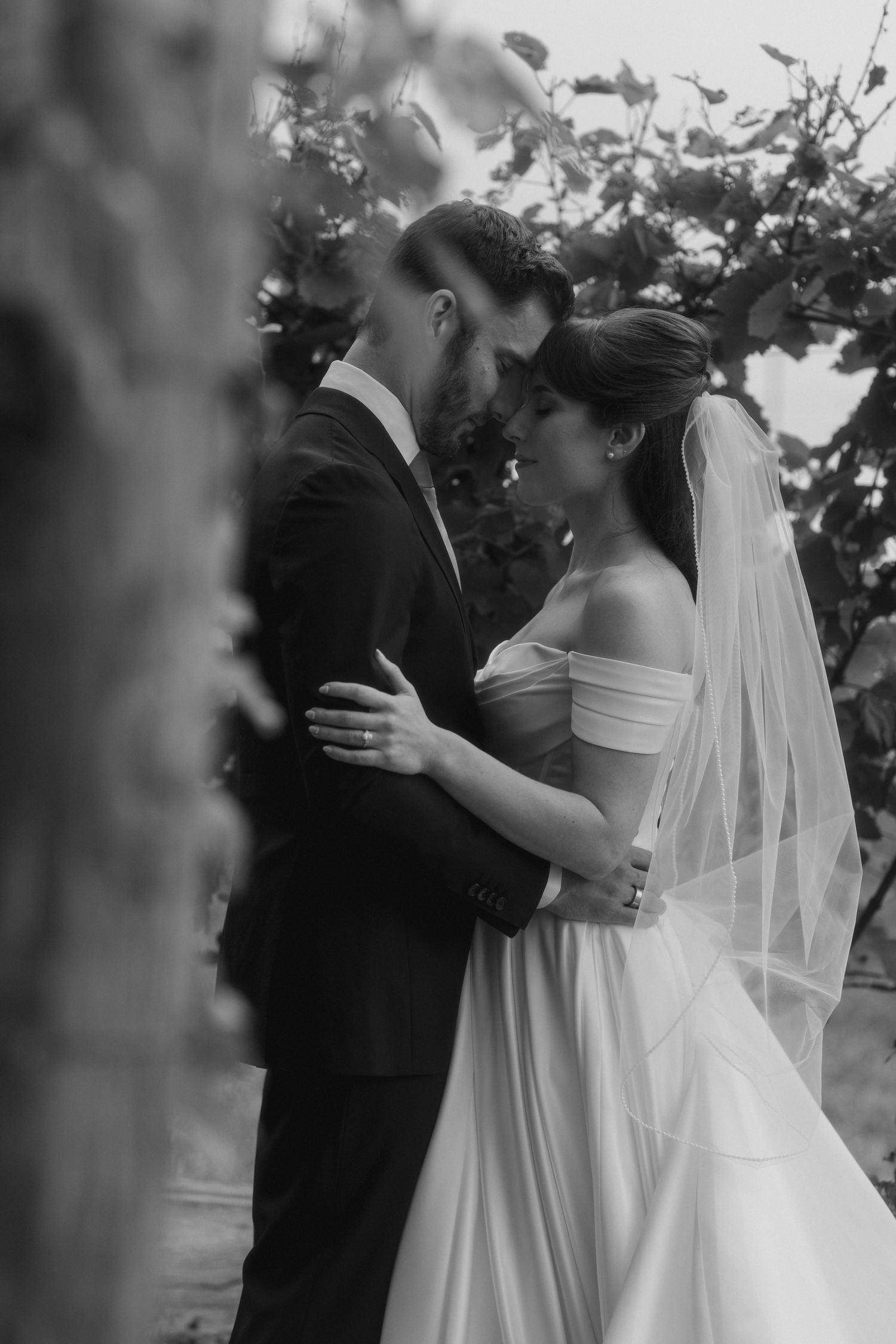 Black and white photo of a bride and groom embracing tenderly among vineyard vines on their wedding day.