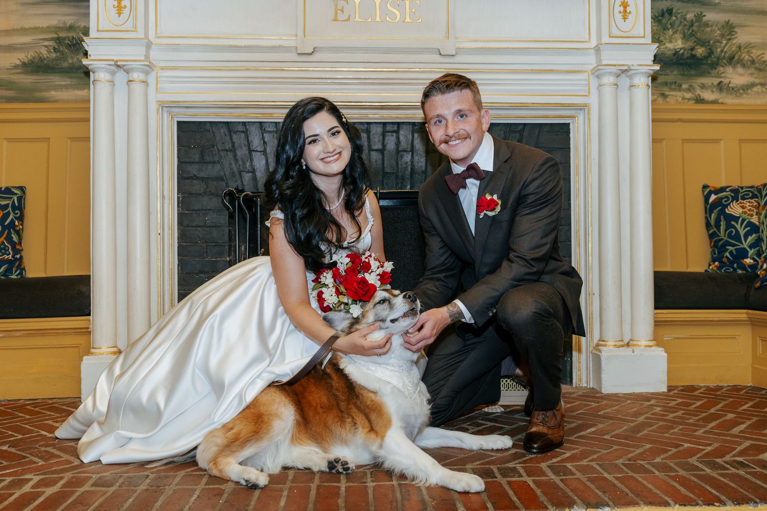 Smiling bride and groom kneel beside their dog in front of elegant fireplace.