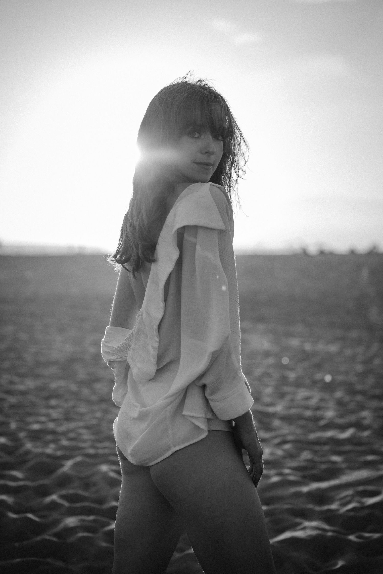 Black and white photo of a woman in an oversized shirt standing on a beach with sun flare behind her.