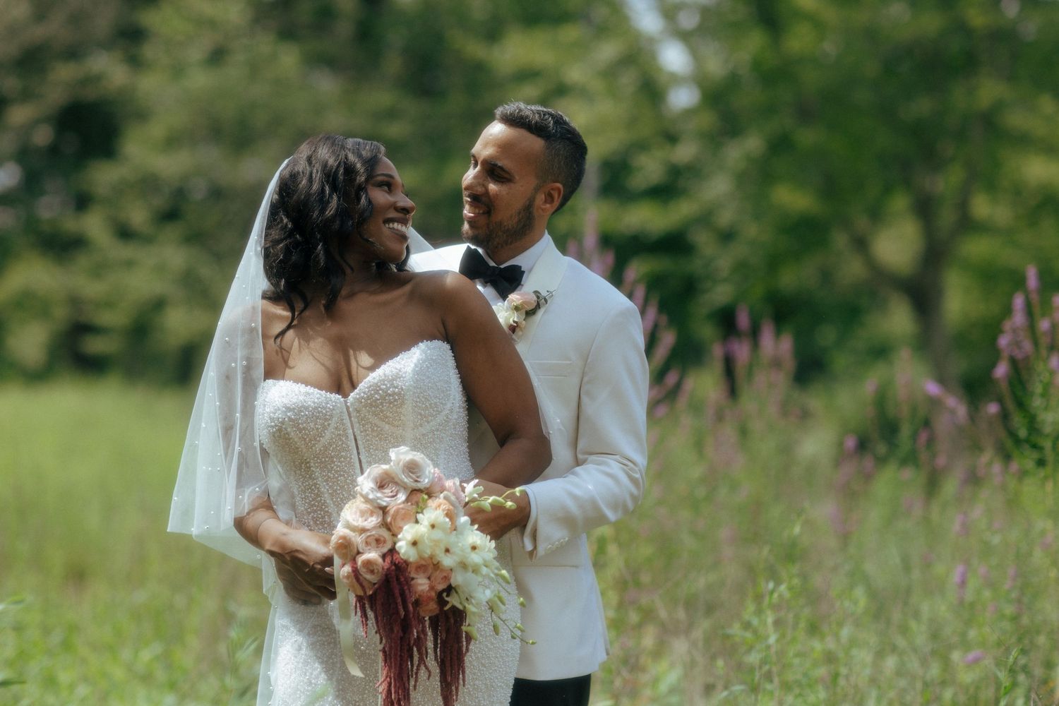 Interracial wedding couple embracing in lush green meadow, bride holding cascading bouquet.