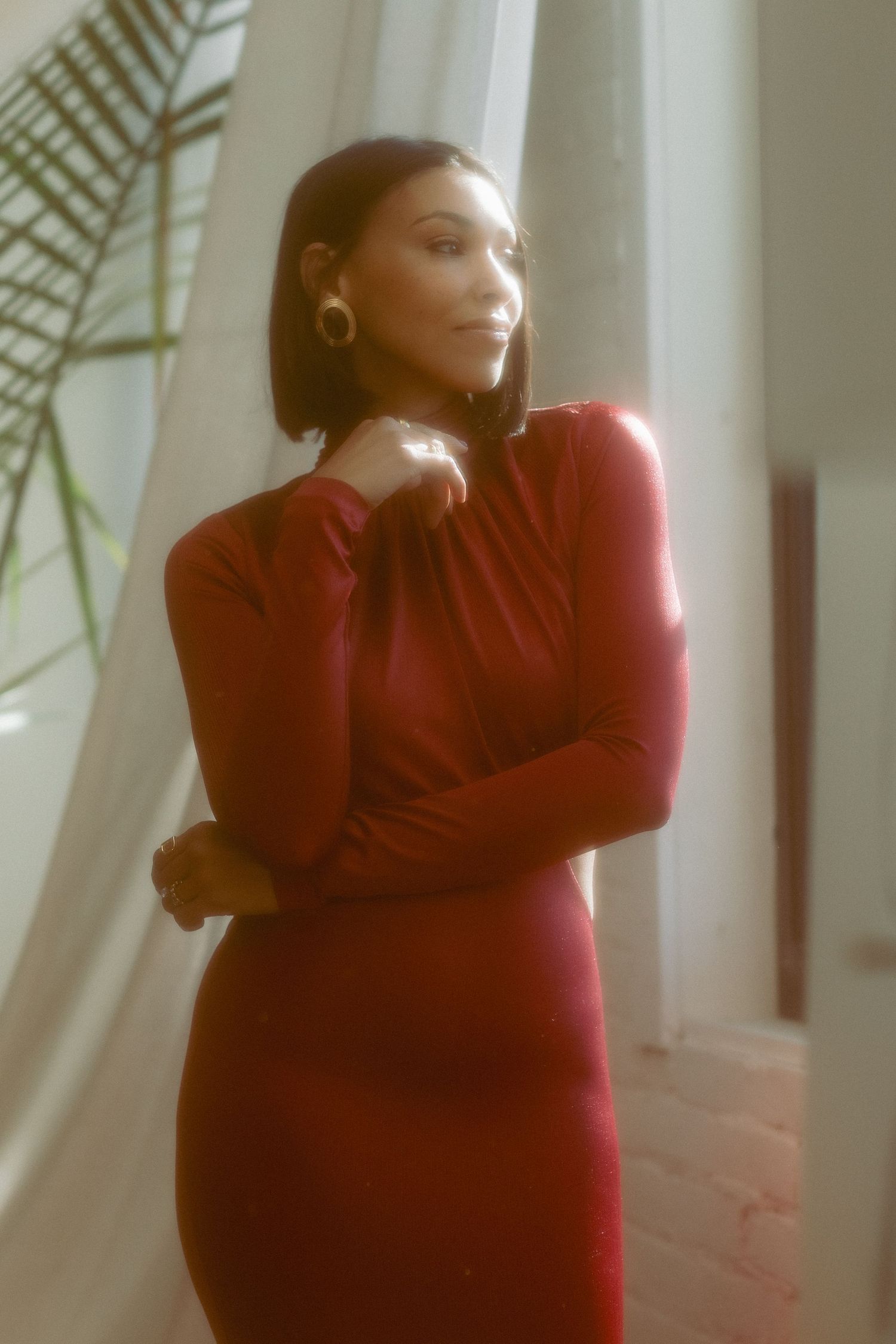 Woman in a red long-sleeve turtleneck dress posing thoughtfully by sheer white curtains in warm cinematic lighting.