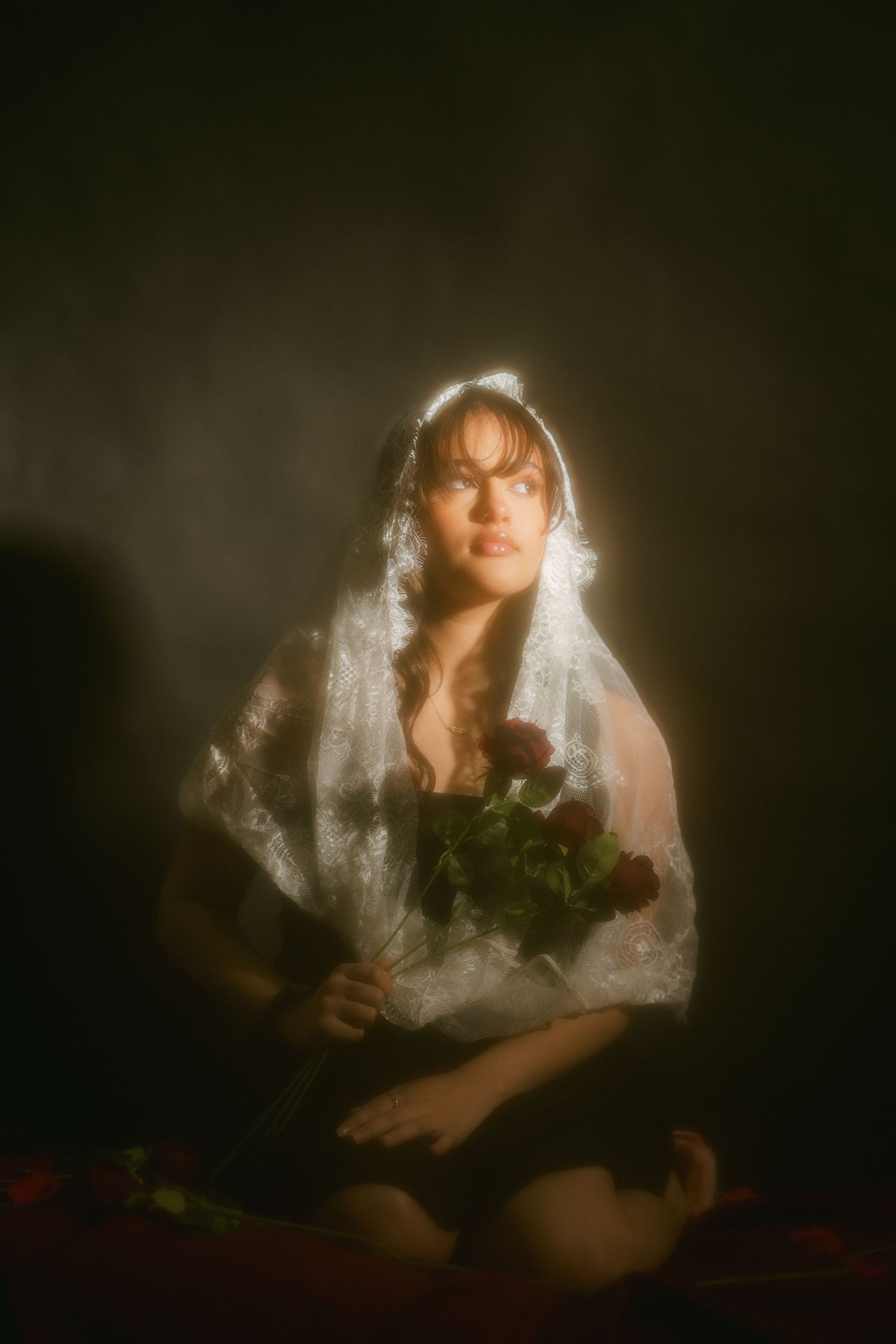 Woman in white lace veil holding bouquet of red roses in dramatic lighting.