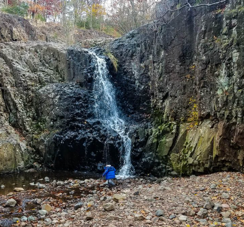 Person in blue jacket sitting by a rocky waterfall surrounded by autumn foliage and dark stone cliffs.