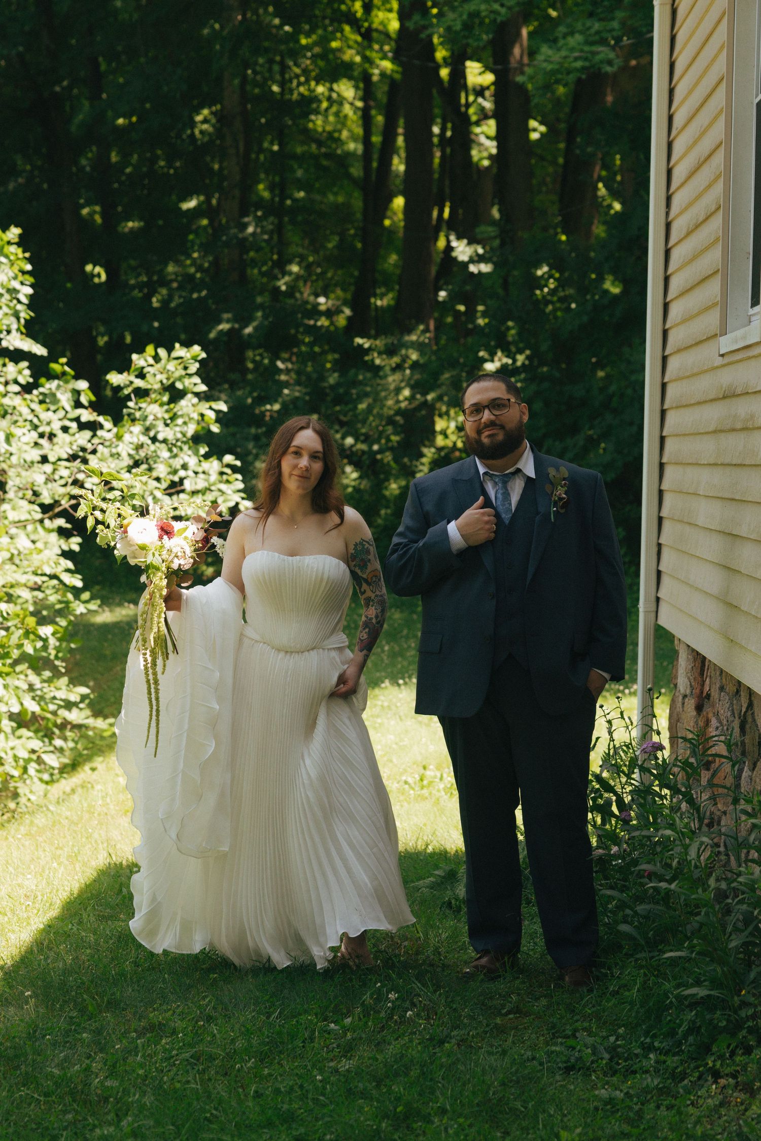 Bride in white gown holding bouquet stands with groom in navy suit outdoors near trees on wedding day.