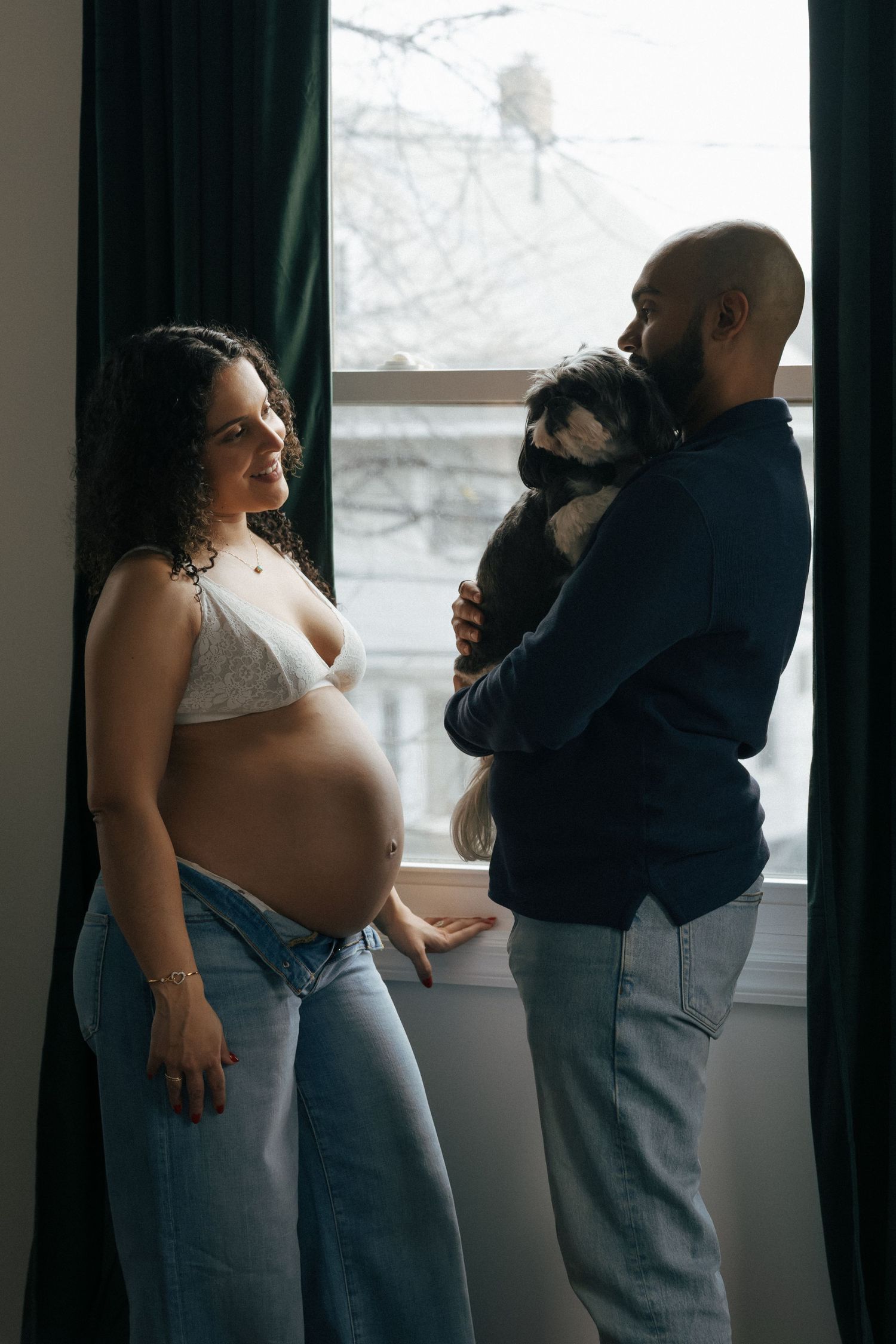 Pregnant woman in white bra and jeans smiles near window as man holds small dog in cozy home setting.