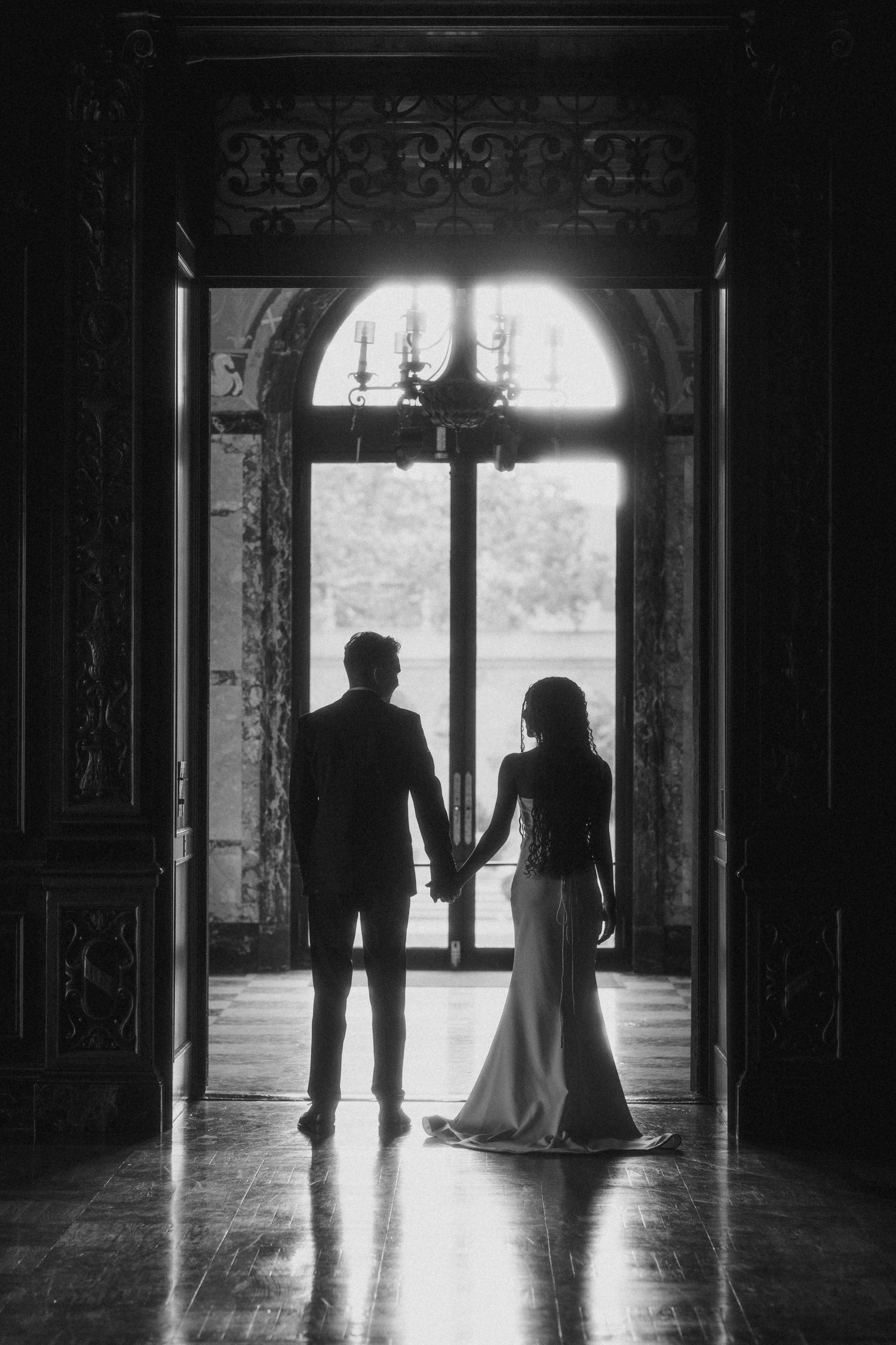 Elegant black and white wedding photo of bride and groom silhouette holding hands before grand arched window in ornate hall.