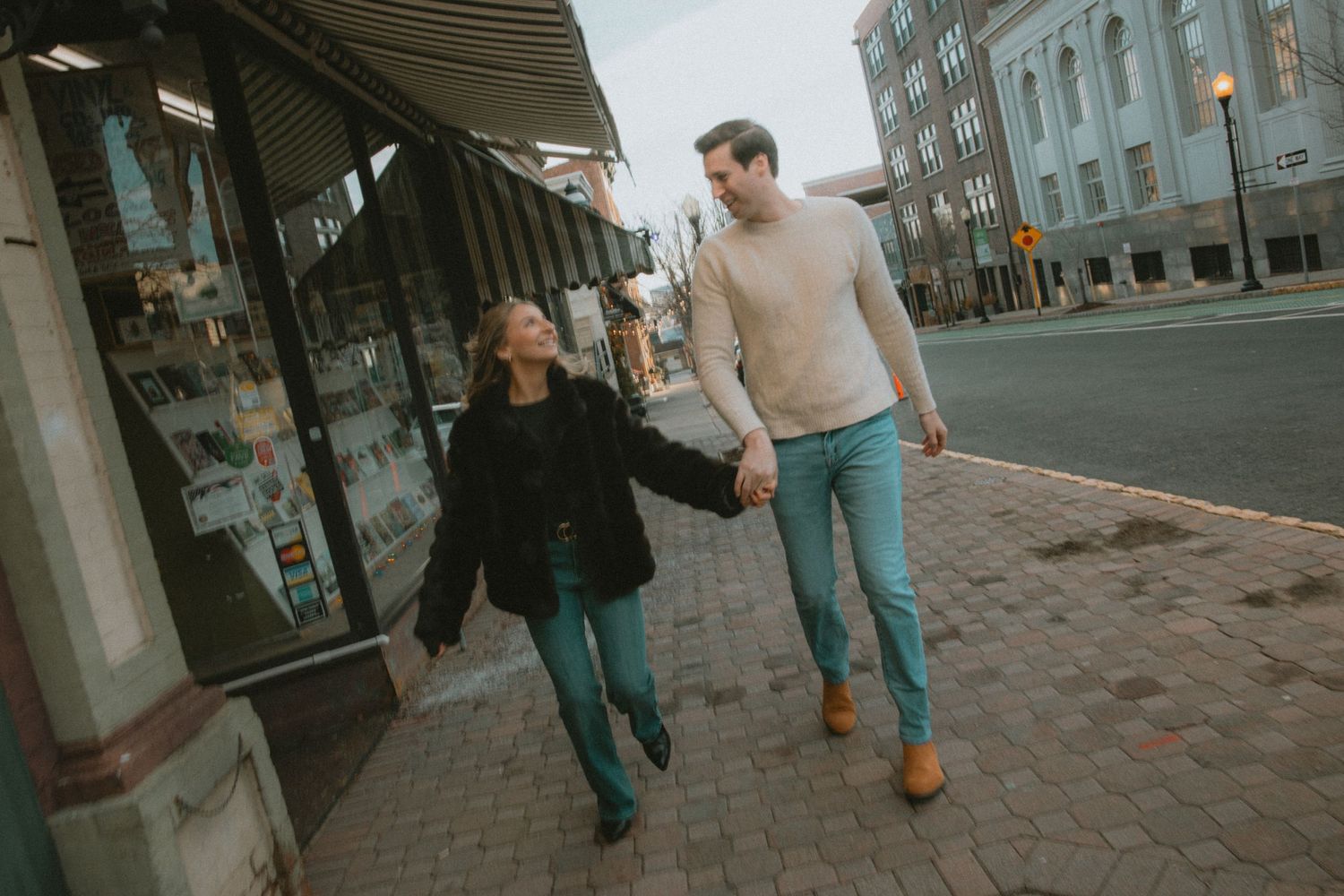 A couple holds hands and walks along a brick sidewalk past a storefront in a charming downtown area.