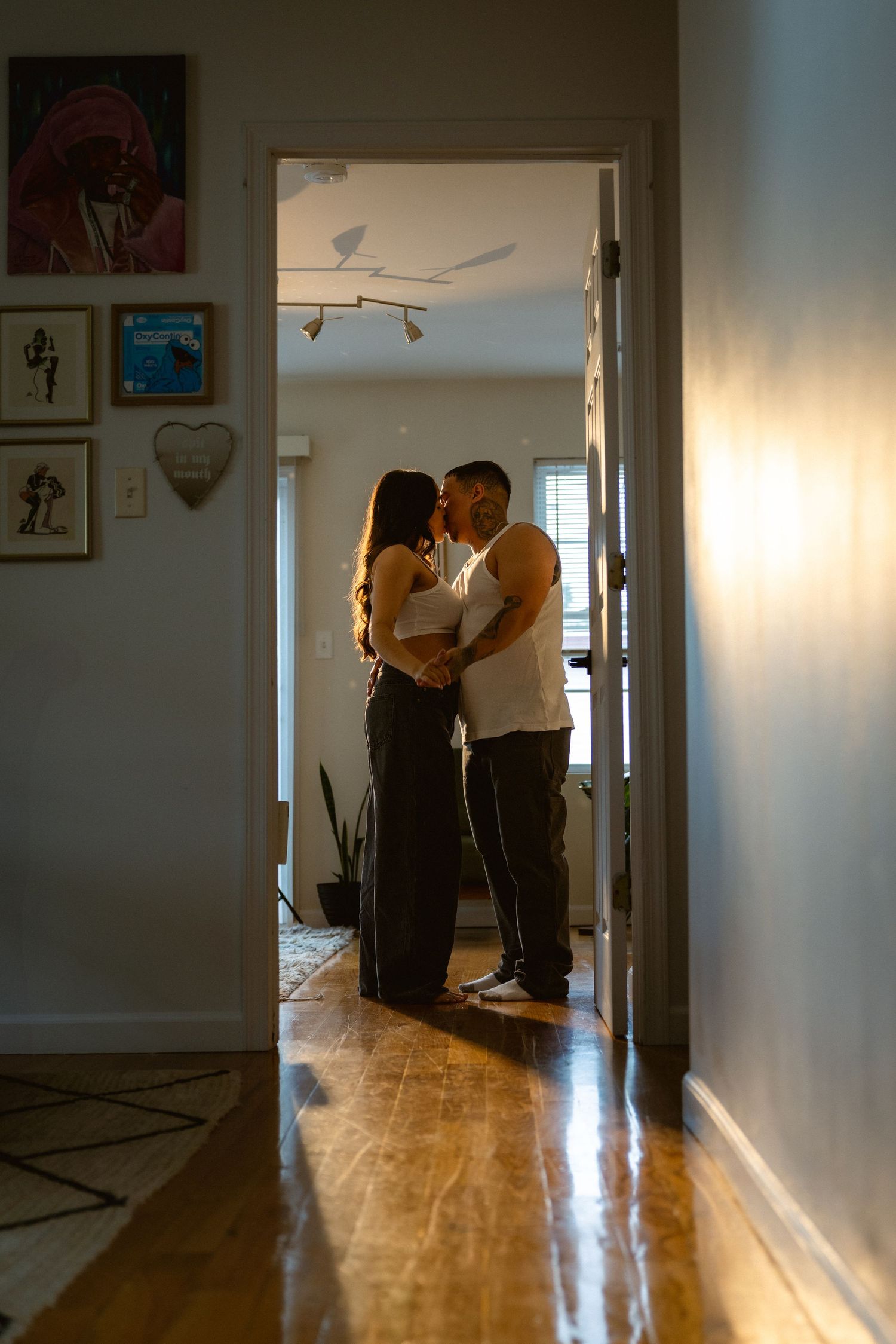 A couple shares an intimate moment kissing in a sunlit doorway of a cozy home with hardwood floors and framed wall art.