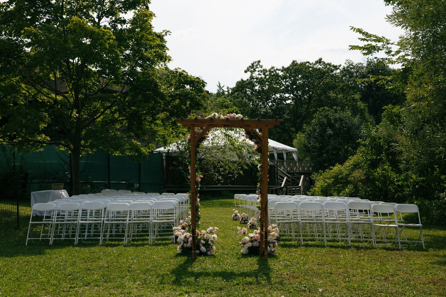 Outdoor wedding ceremony setup with floral arch and white chairs on green lawn.