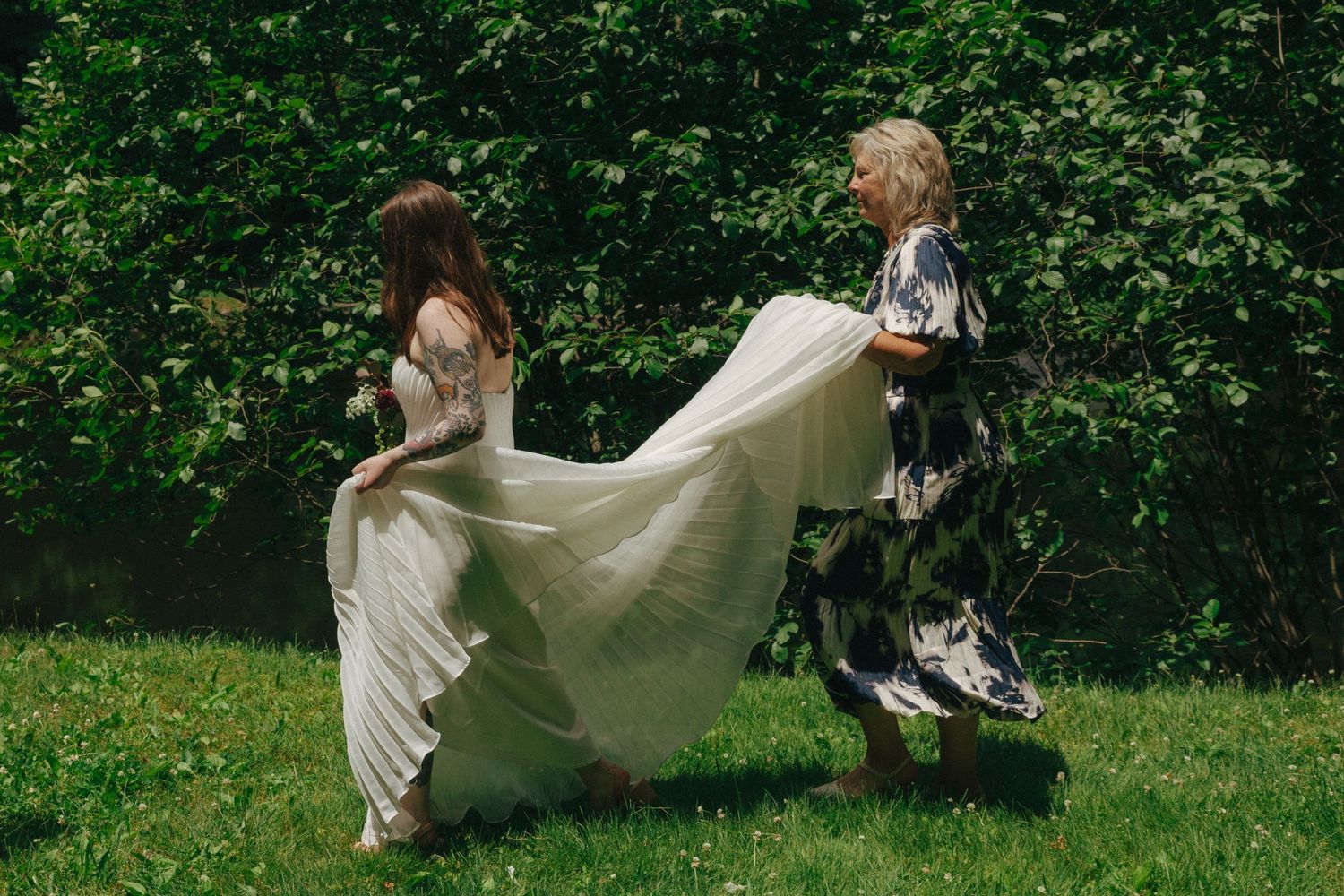 Bride in white lace gown has train adjusted by guest in floral dress on green lawn.