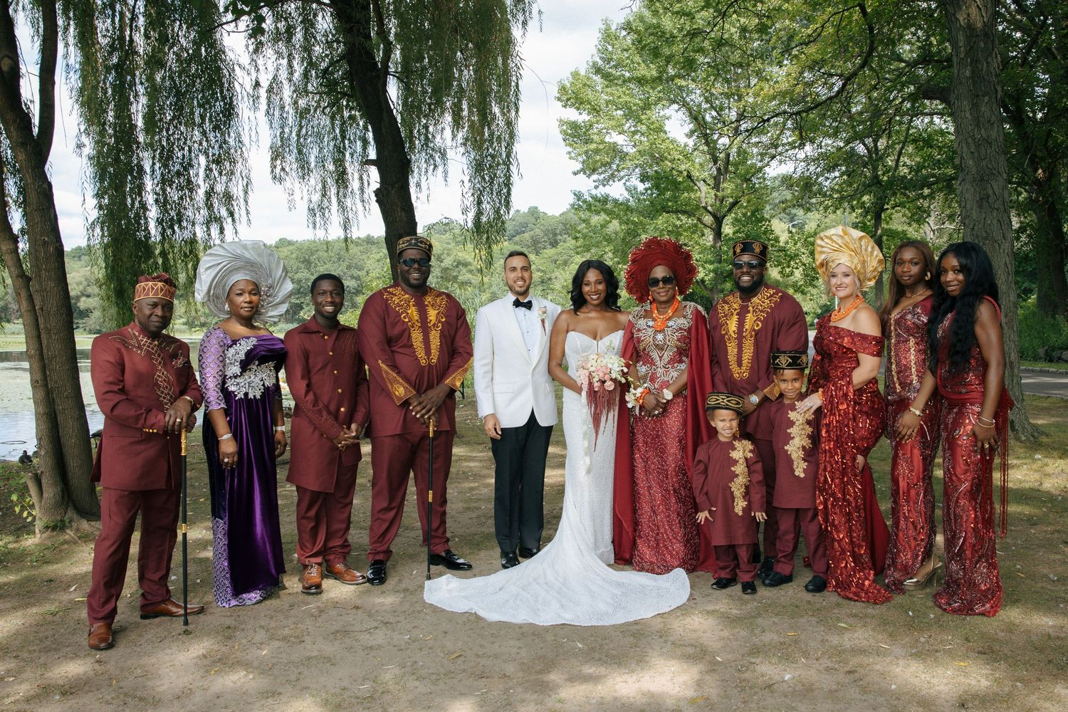 Wedding party in matching burgundy African attire poses outdoors with bride and groom in white.