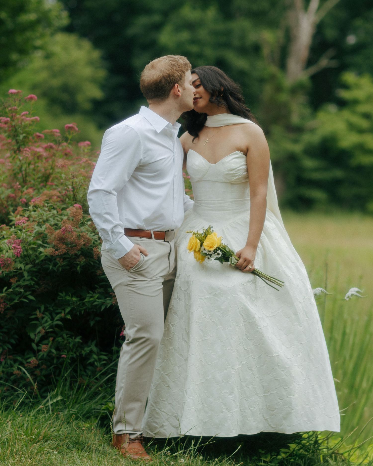 A bride and groom share a kiss outdoors, she holds yellow flowers in a white gown, surrounded by lush greenery.