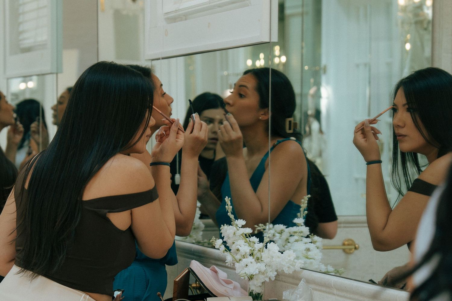Women applying makeup in front of a large mirror, preparing for a special event in an elegant setting.