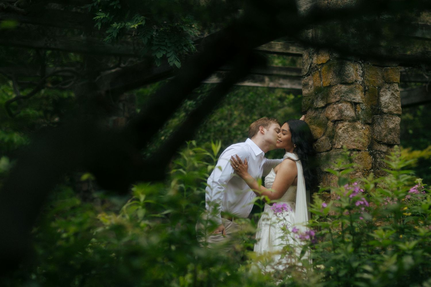 Couple sharing a romantic kiss among lush green garden foliage beside a stone wall.