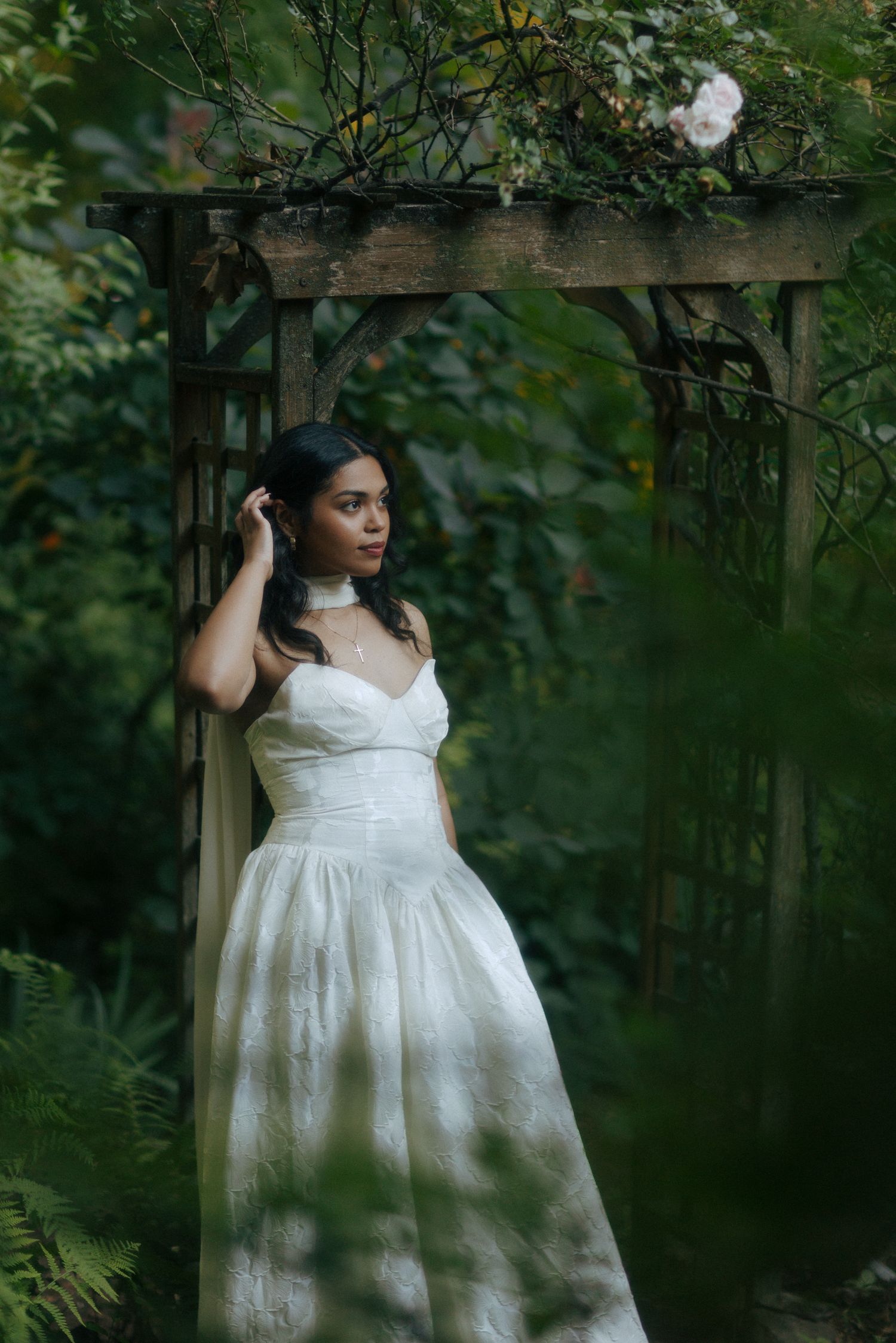 Woman in white gown poses under wooden garden arbor surrounded by lush green foliage and roses.