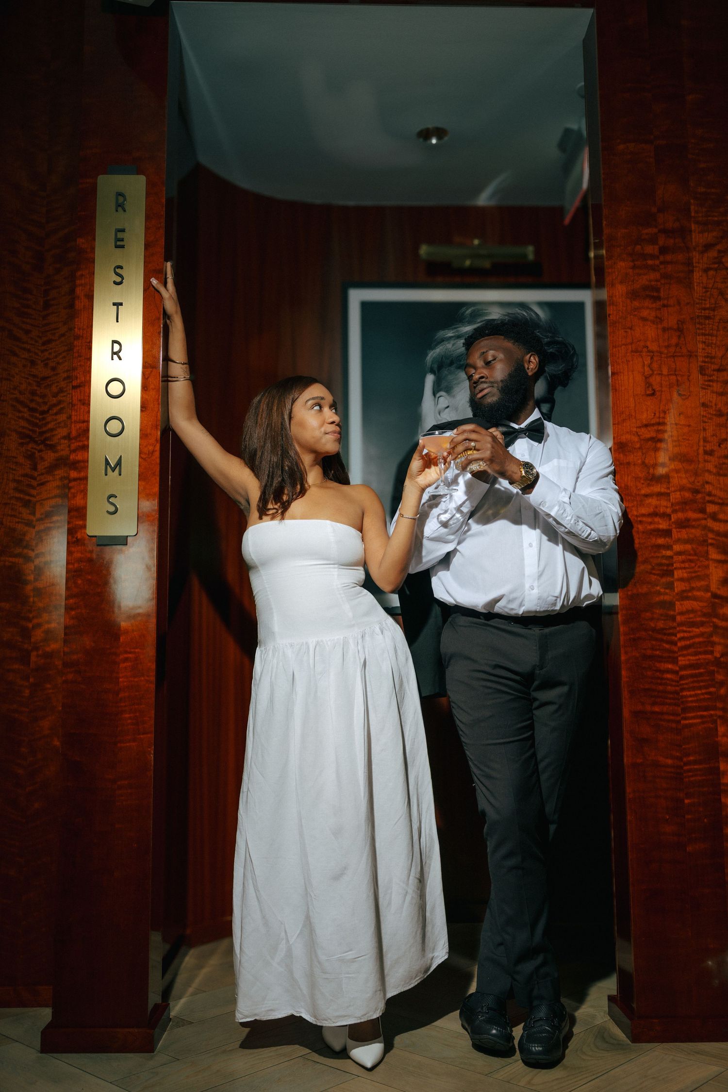 Fashionable Black couple in formal wear posing together near a restrooms sign in an elegant venue.