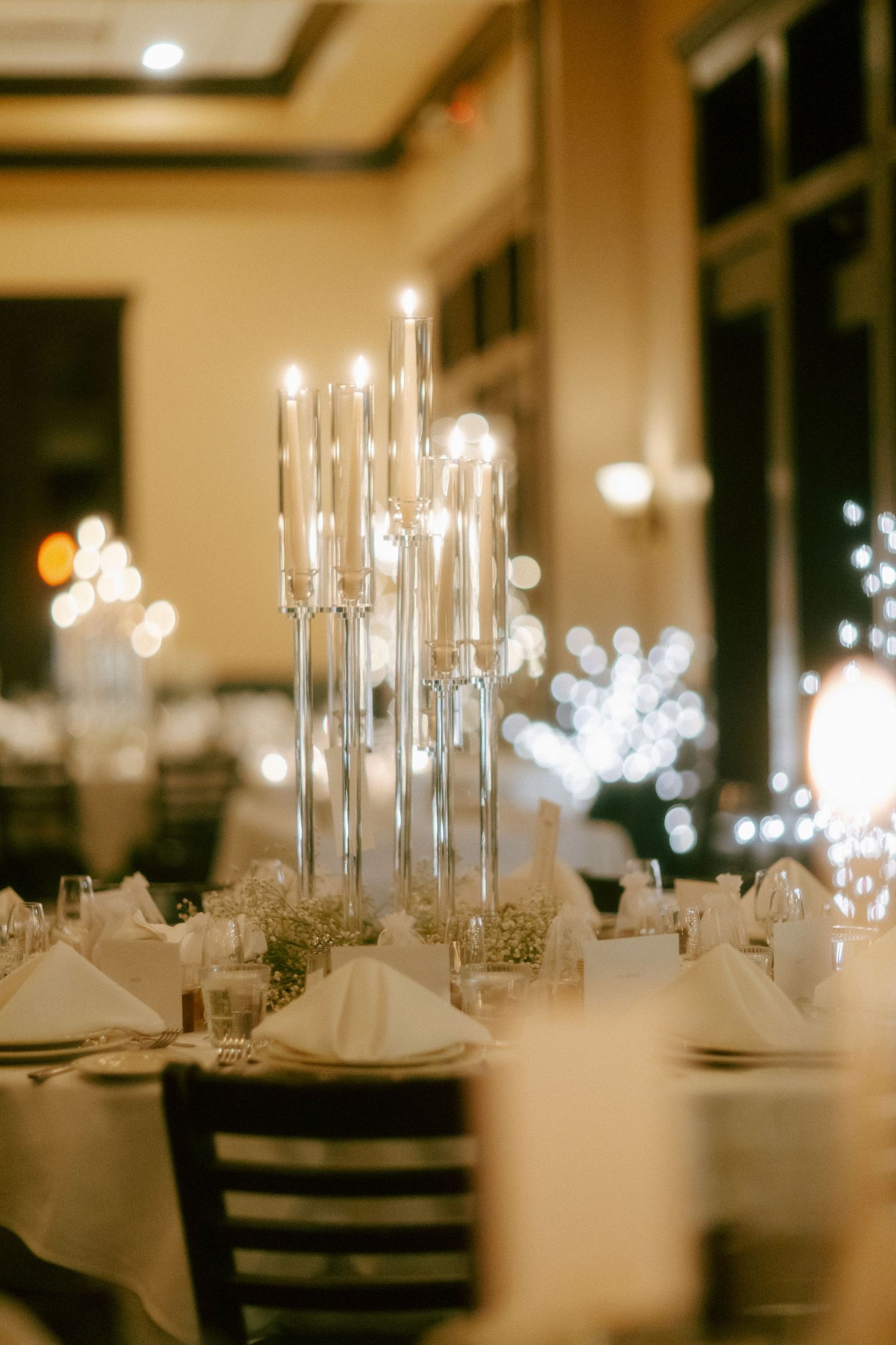 Elegant wedding reception table with tall silver candelabra centerpiece and white folded napkins in a warmly lit ballroom.