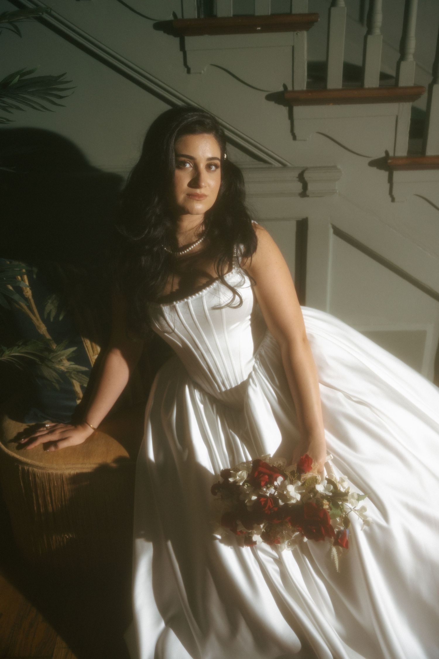 Elegant bride in white corset ballgown holds red and white bouquet while seated near sunlit staircase.