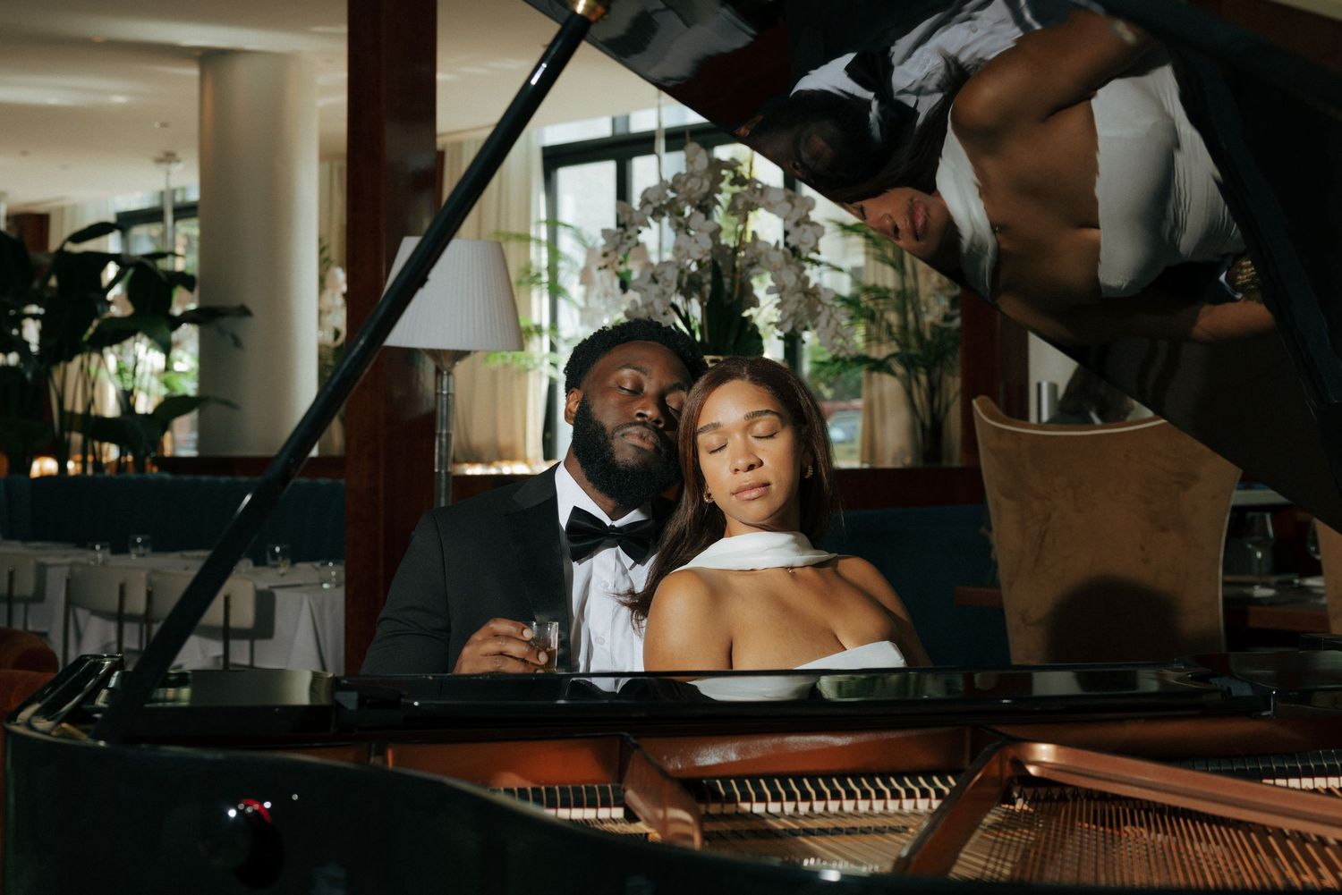 An elegant couple poses at a grand piano, man in tuxedo and woman in strapless gown in a luxury venue.