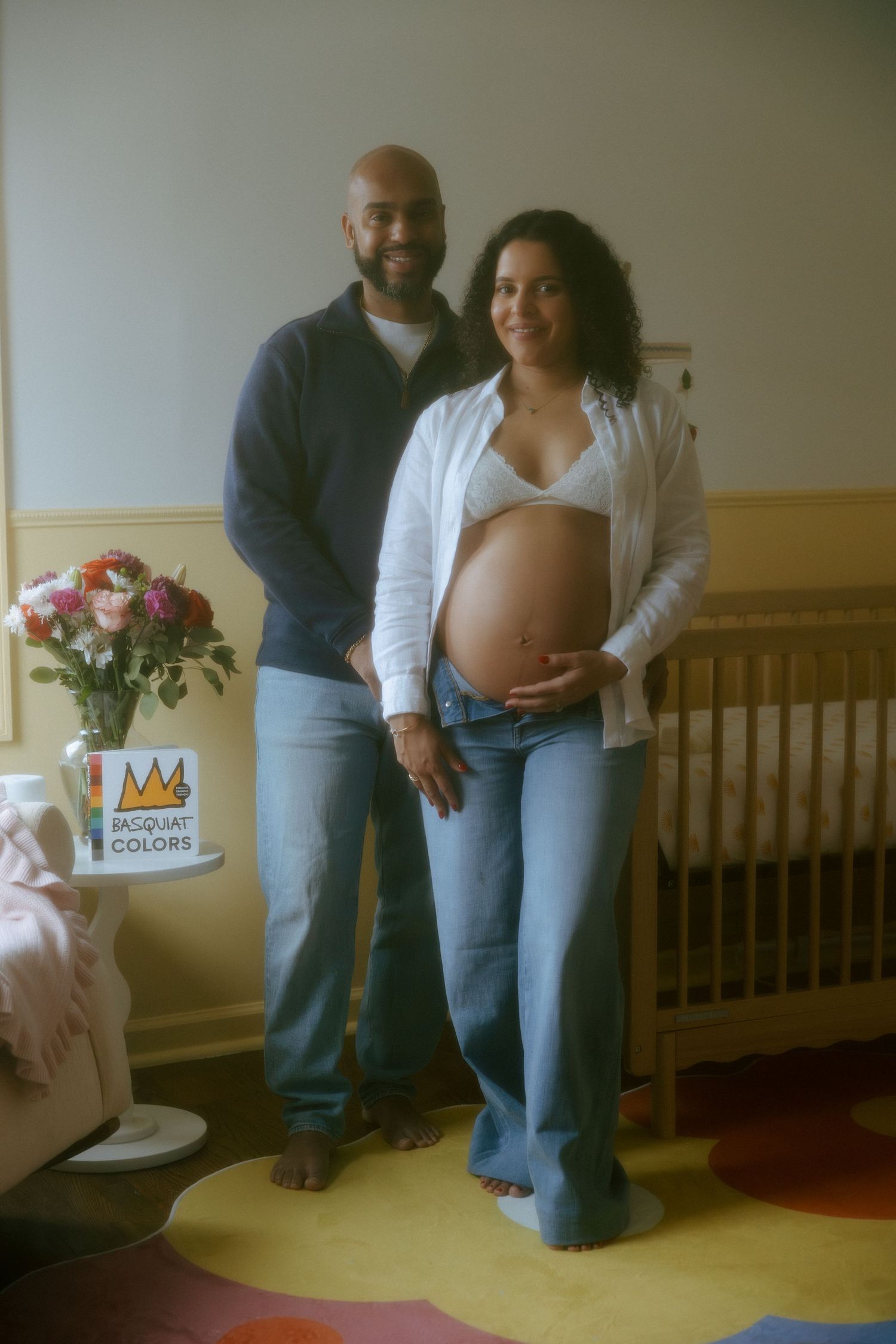 Pregnant woman in white bralette and open shirt poses with partner in nursery room with crib and flowers.