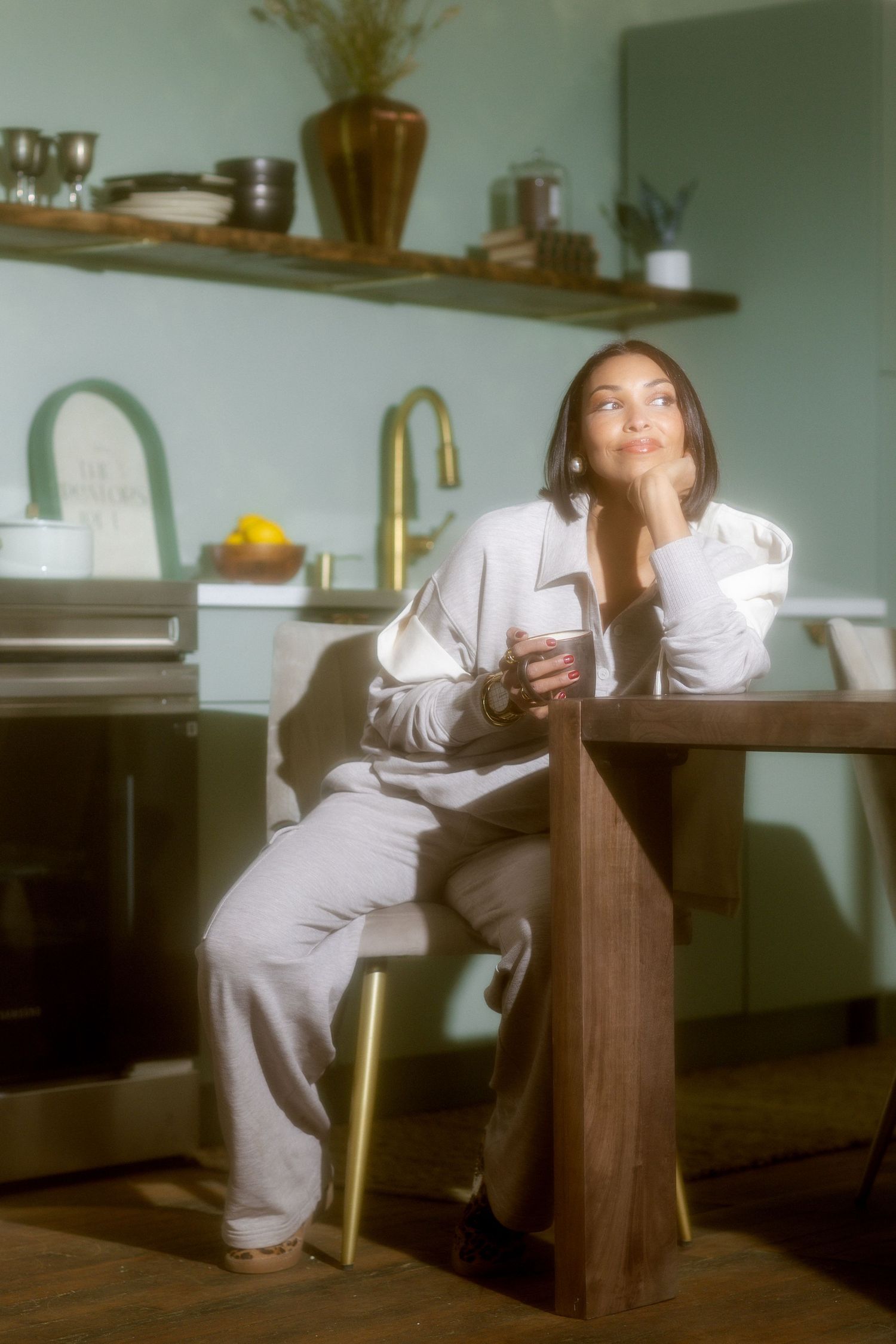 Woman in gray outfit sitting at kitchen island, gazing upward thoughtfully in a sunlit modern kitchen.