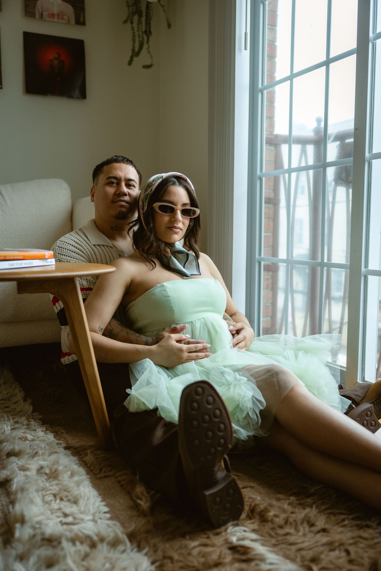 Pregnant woman in mint green tulle dress with sunglasses sits on floor leaning against man in cozy living room.