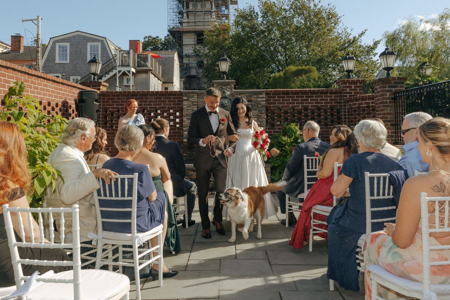 Outdoor rooftop wedding ceremony with guests seated on white chairs as couple exchanges vows with dog present.
