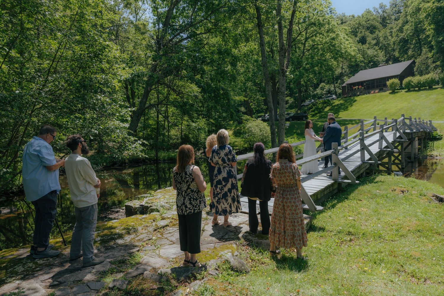 Group of people standing near a wooden bridge surrounded by lush green trees and a rustic cabin in a scenic outdoor setting.