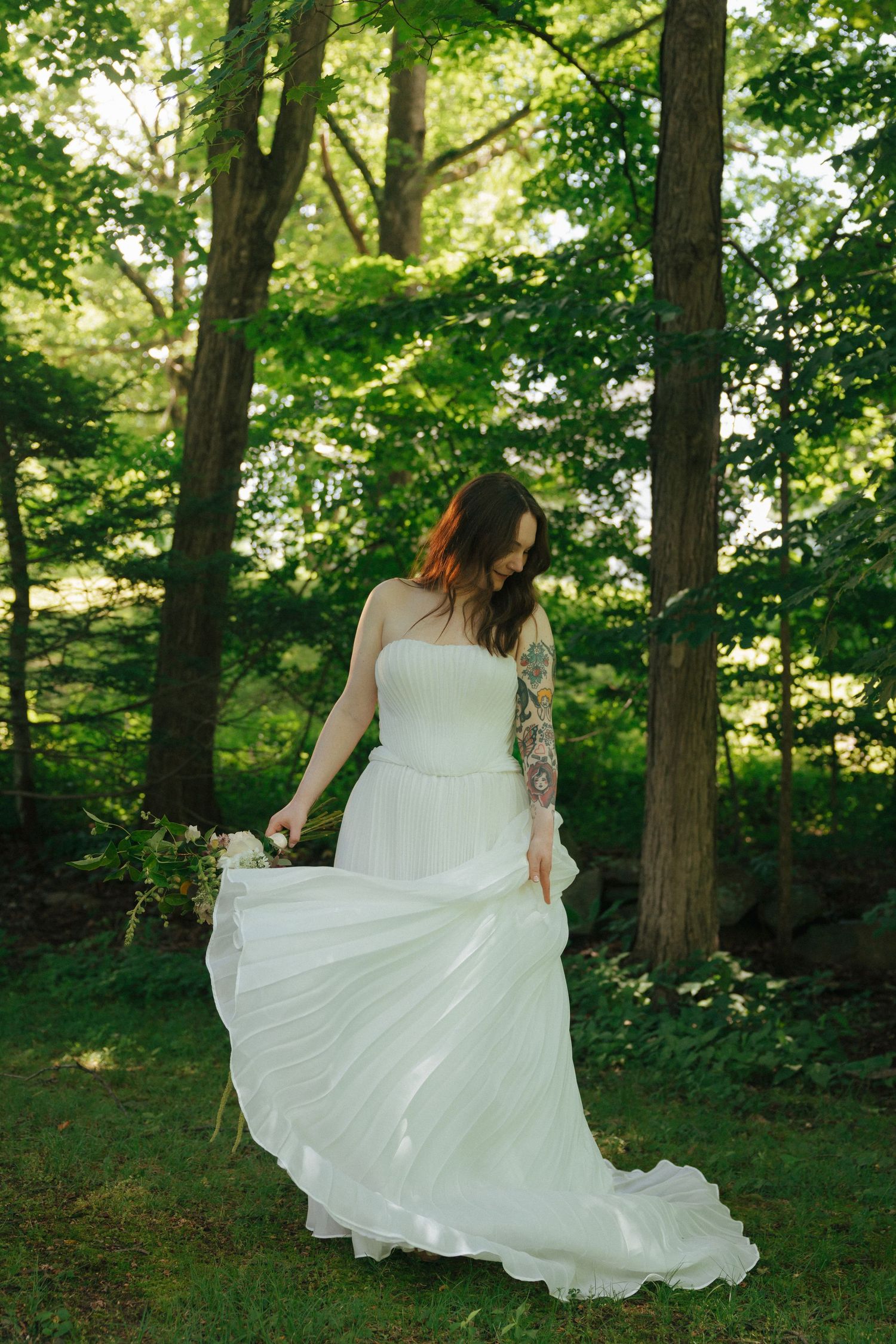 Bride in strapless white gown with floral tattoo sleeve holds bouquet in lush green forest setting.