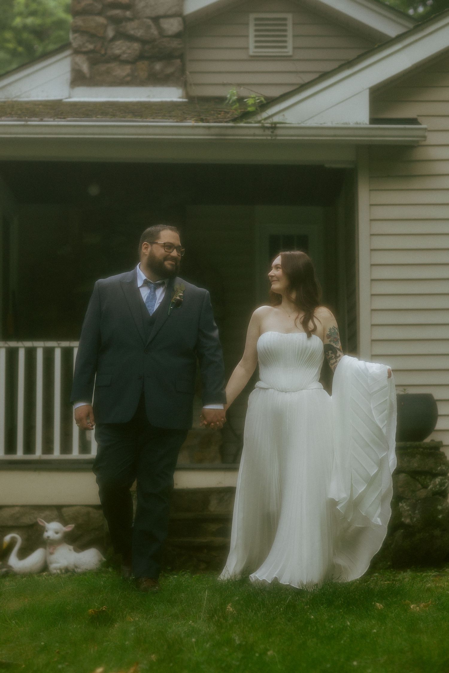 Bride and groom hold hands smiling outside a charming house, bride in white gown, groom in navy suit.