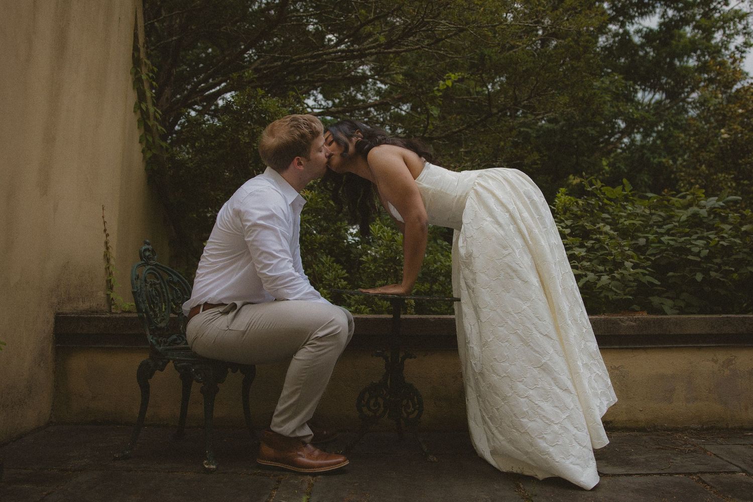 A bride in a white gown leans in to kiss her groom seated on a bench outdoors at a garden wedding venue.