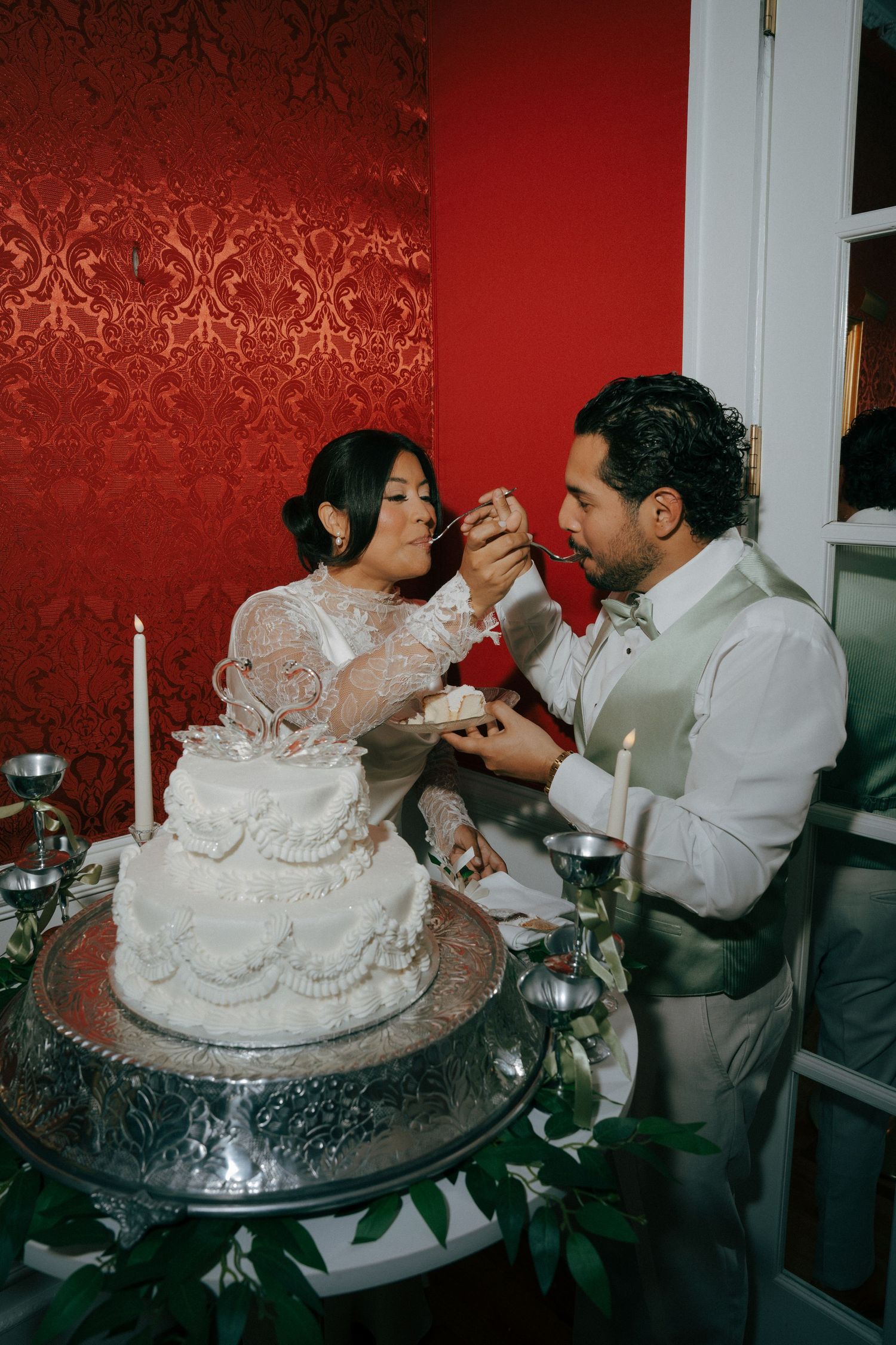 Bride and groom feeding each other wedding cake at elegant reception with red damask walls and white tiered cake.