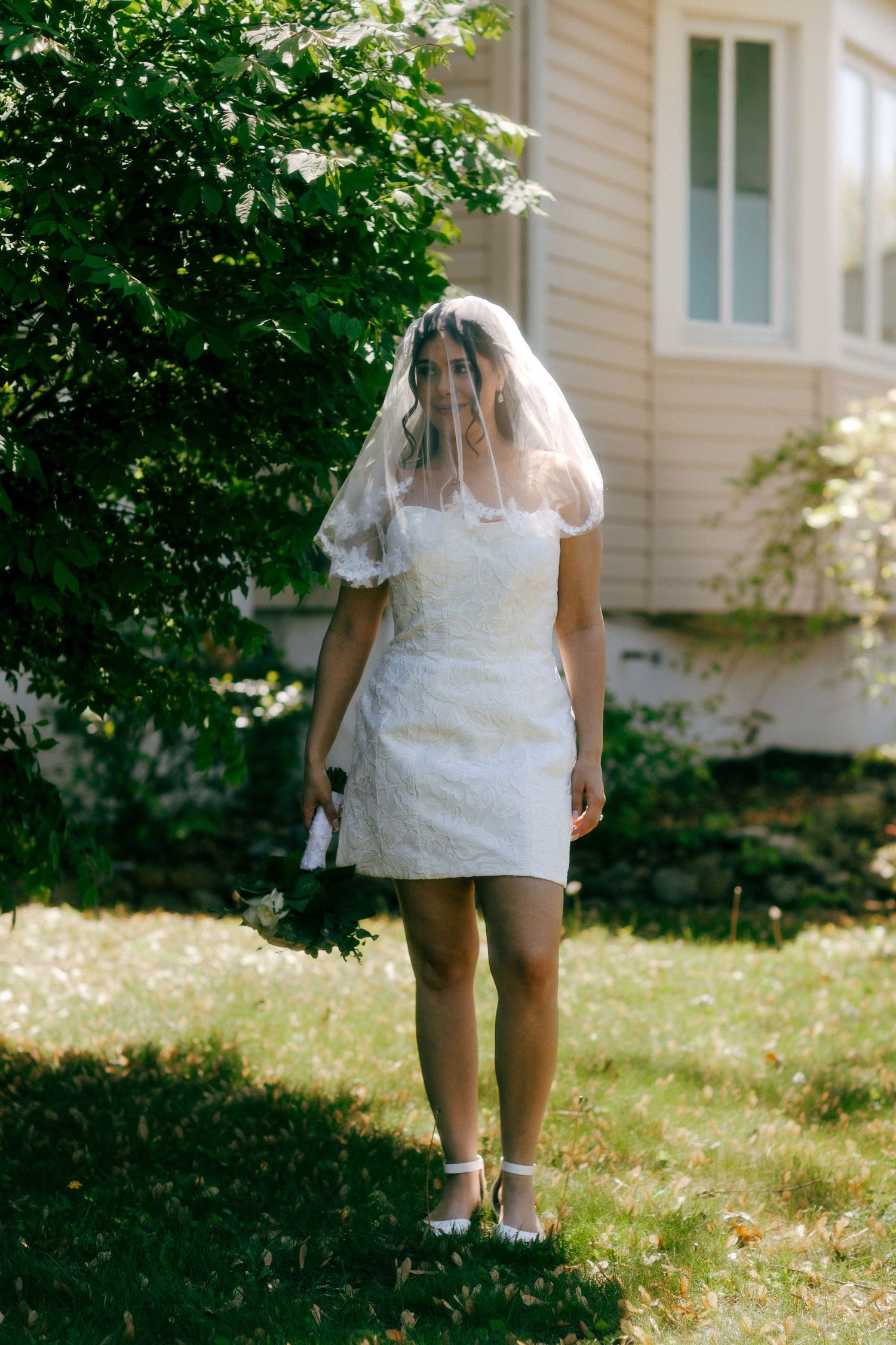 Bride in short white lace dress and veil standing on green lawn near bushes outside a house.