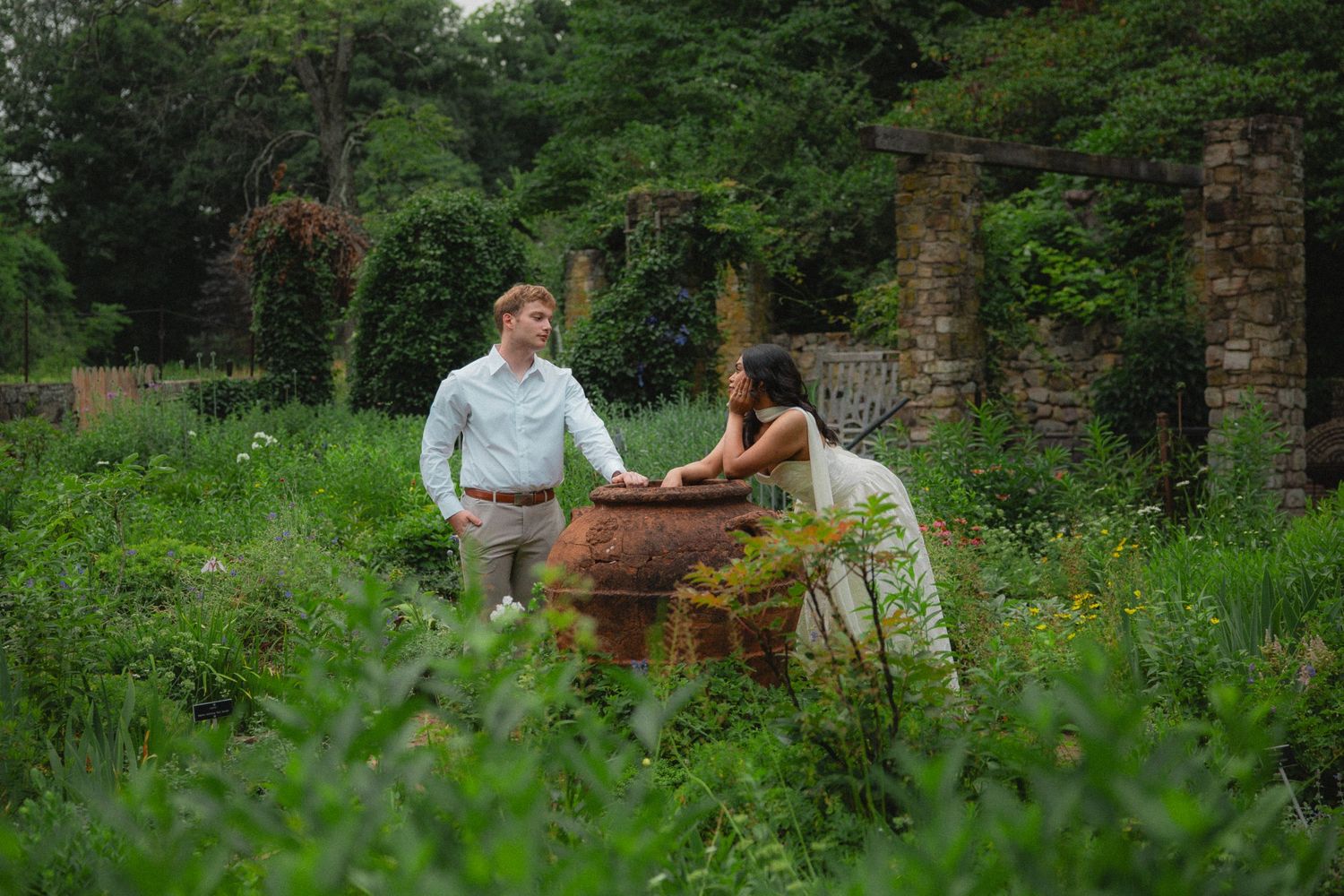 Couple standing together in a lush green garden beside a large terracotta planter with stone ruins in background.